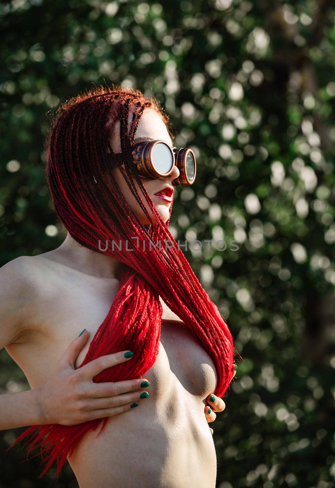 Semi-nude glamorous girl with scarlet dreadlocks and welding glasses closes the breast with scarlet dreadlocks
