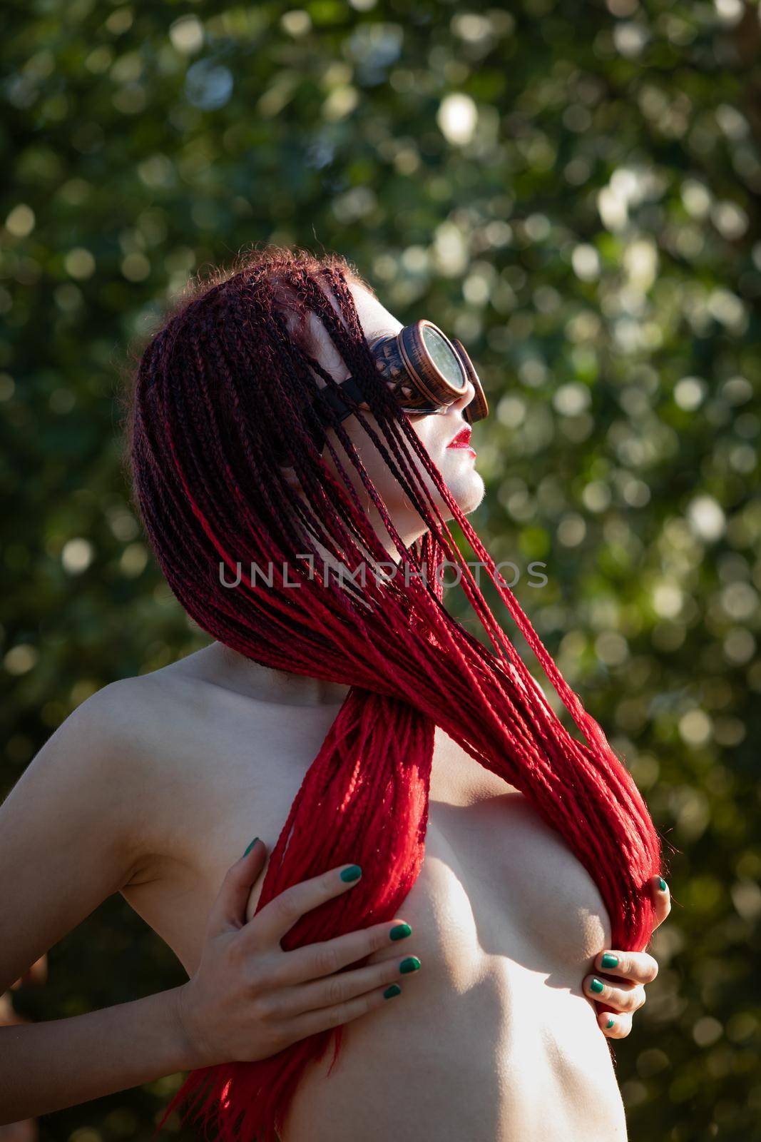 Semi-nude glamorous girl with scarlet dreadlocks and welding glasses closes the breast with scarlet dreadlocks