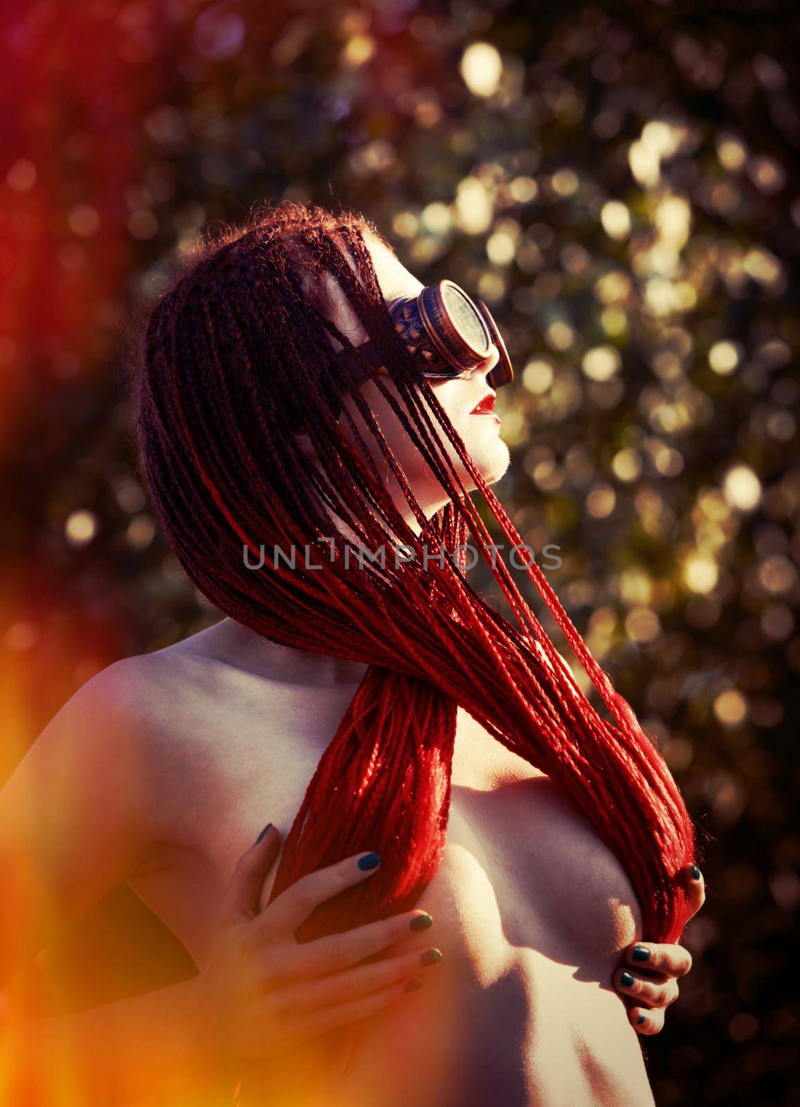 Semi-nude glamorous girl with scarlet dreadlocks and welding glasses closes the breast with scarlet dreadlocks