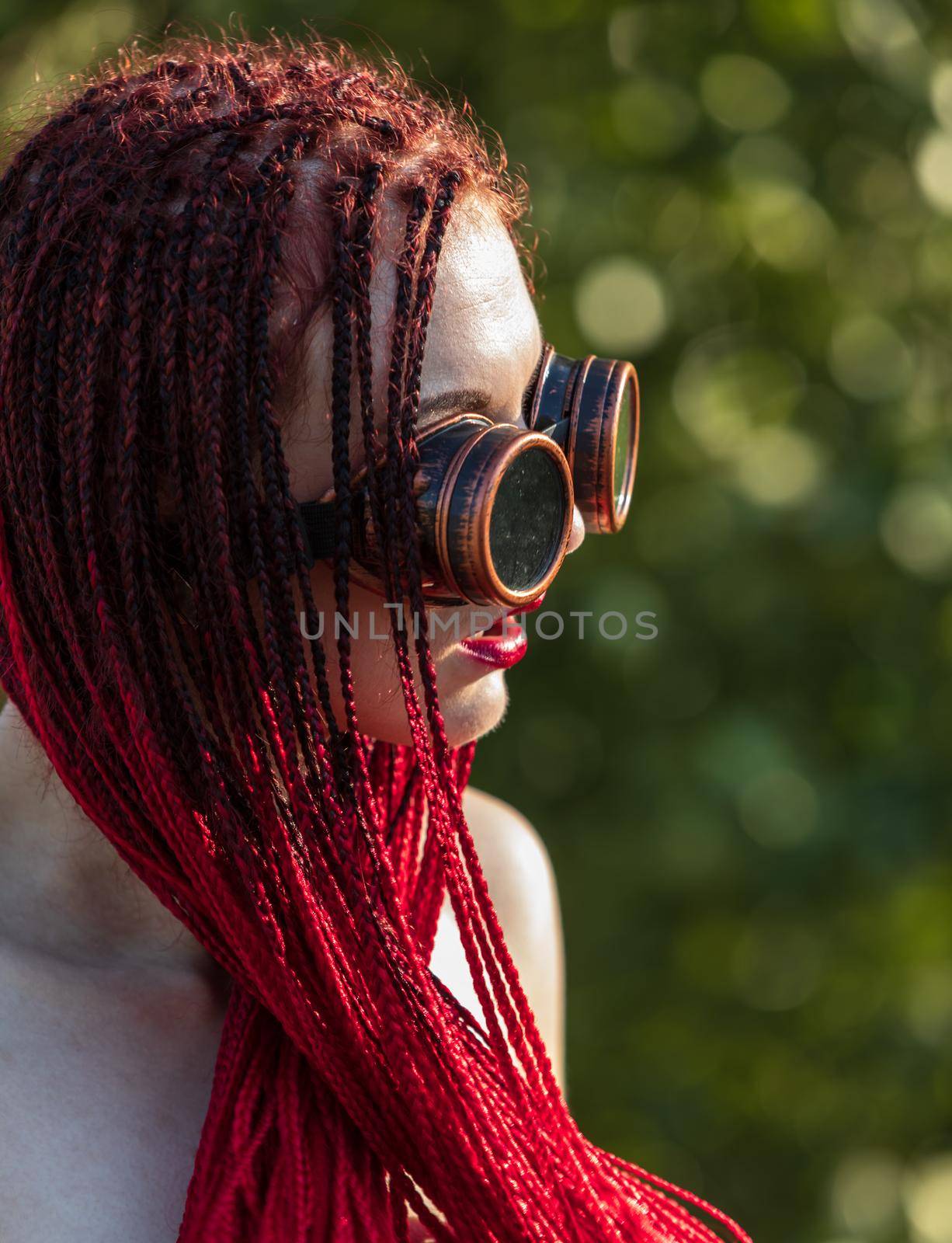 Semi-nude glamorous girl with scarlet dreadlocks and welding glasses closes the breast with scarlet dreadlocks