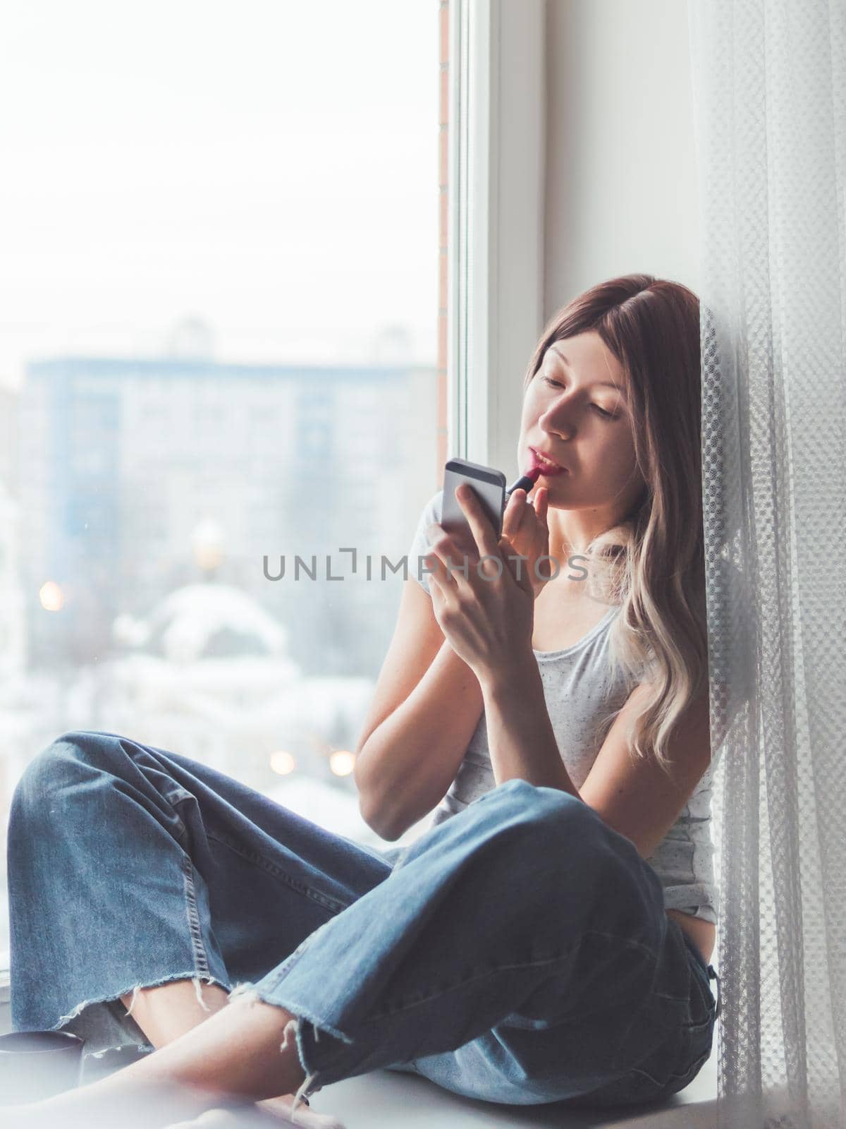 Woman with curly hair paints her lips with lipstick and uses smartphone as mirror. Fast make-up. Morning routine. Cup of hot coffee on windowsill.