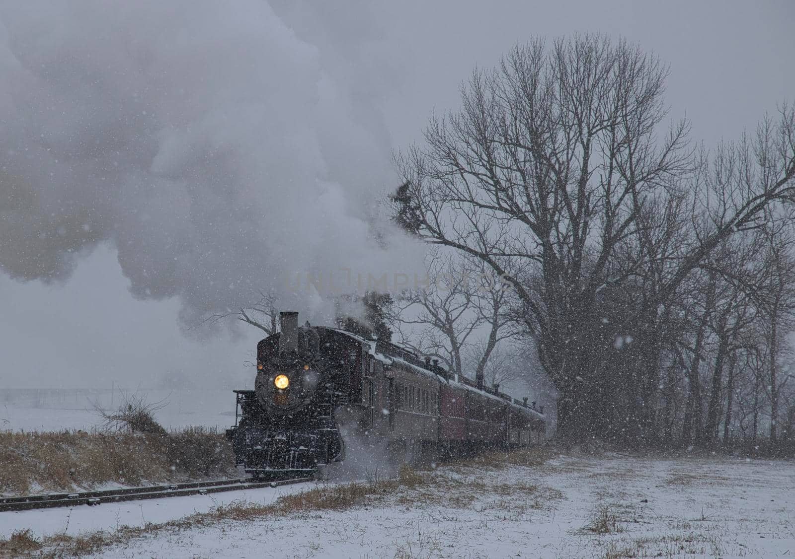 View of An Antique Restored Steam Locomotive Blowing Smoke and Steam Traveling Thru Farmlands and Countryside in a Snow Storm