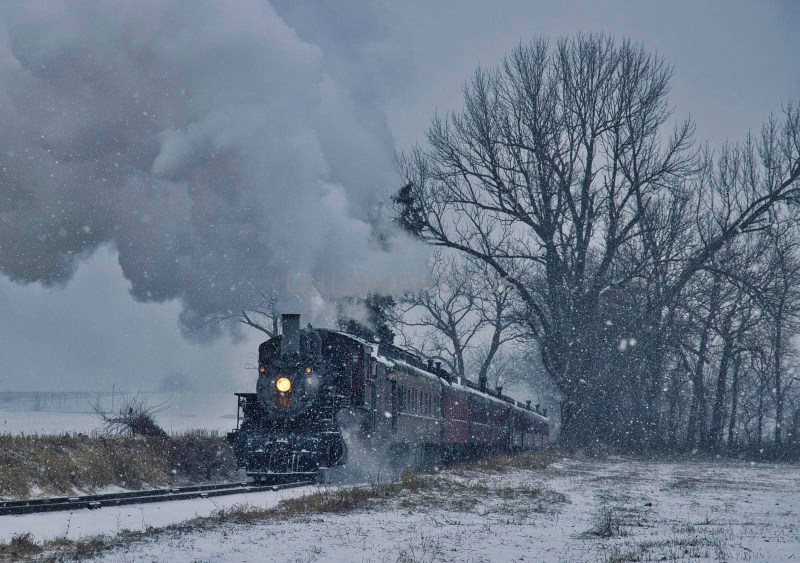 View of An Antique Restored Steam Locomotive Blowing Smoke and Steam Traveling Thru Farmlands and Countryside in a Snow Storm