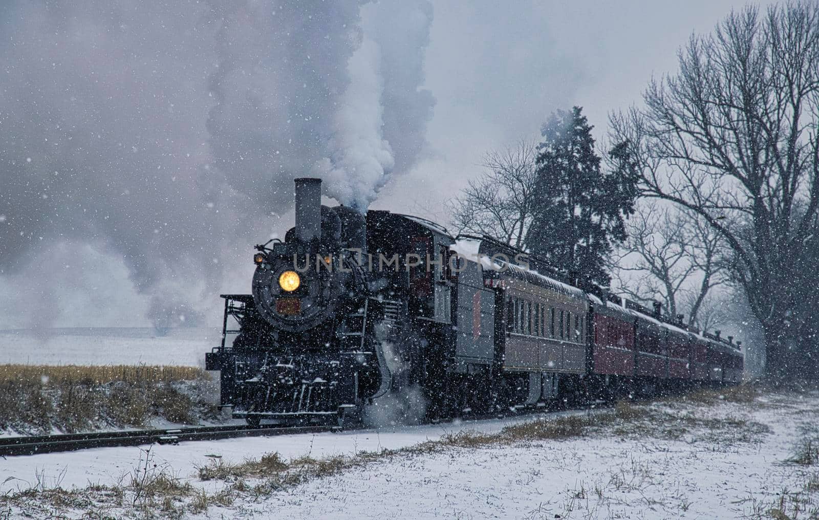View of An Antique Restored Steam Locomotive Blowing Smoke and Steam Traveling Thru Farmlands and Countryside in a Snow Storm