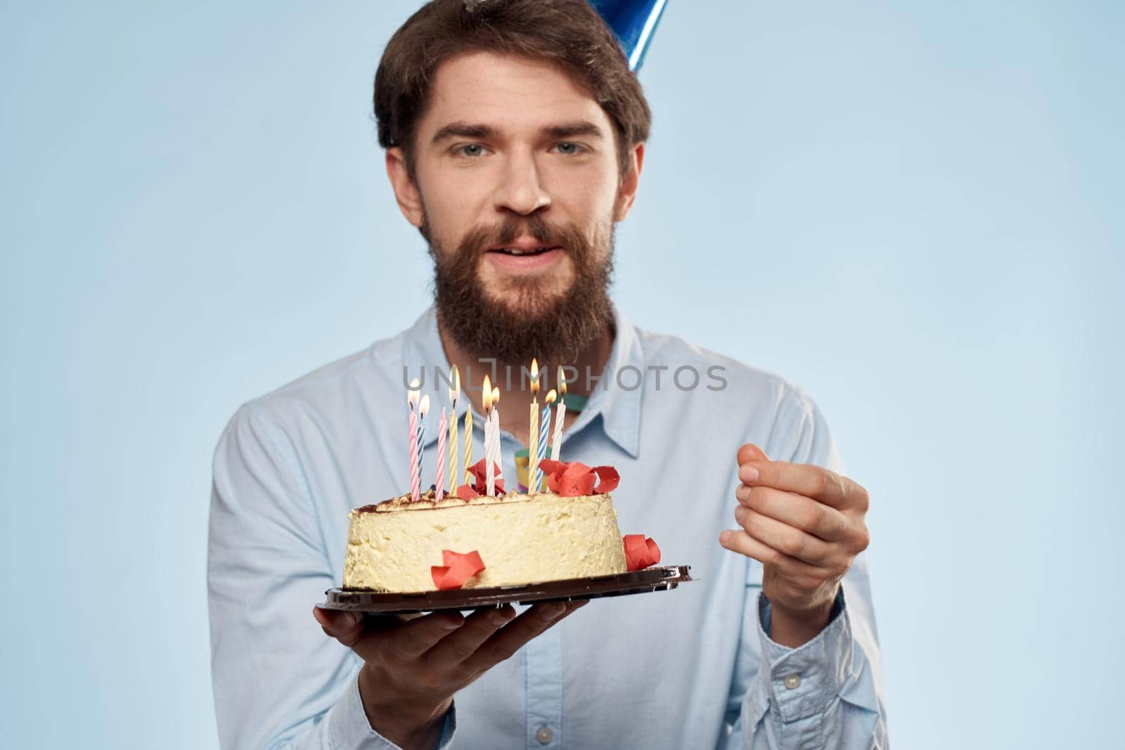 Bearded man with a plate of cake on a blue background and a birthday party hat on his head. High quality photo