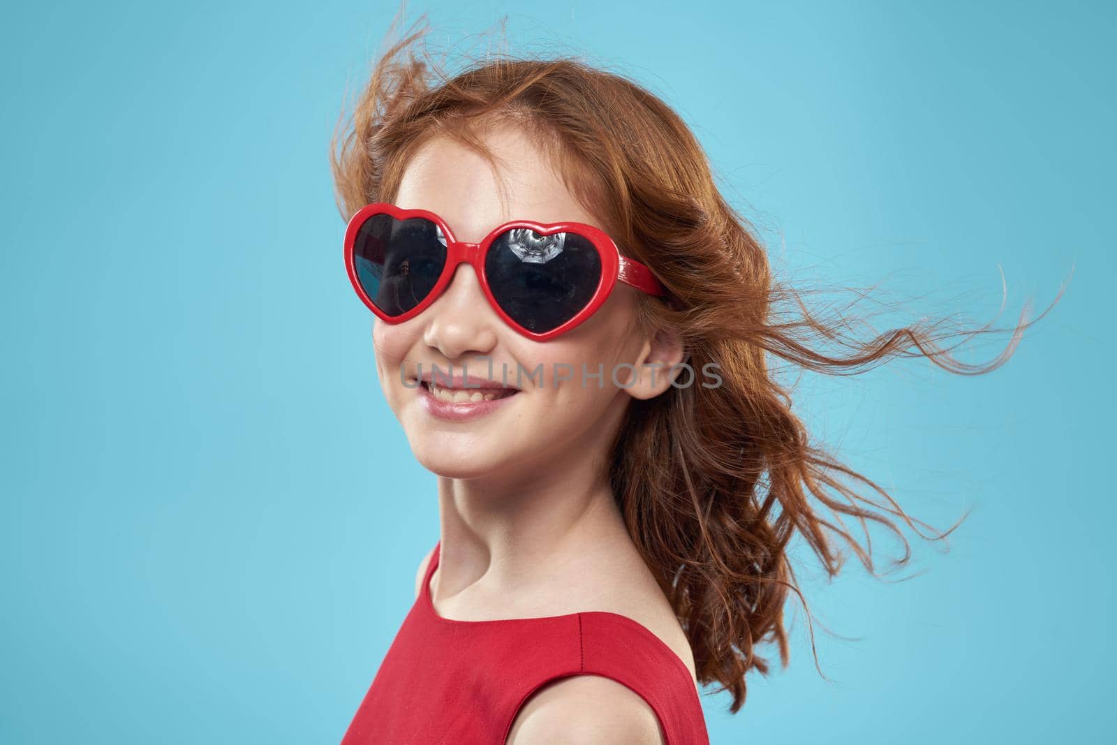 Little girl in sunglasses in the form of hearts red dress curly hair blue background. High quality photo