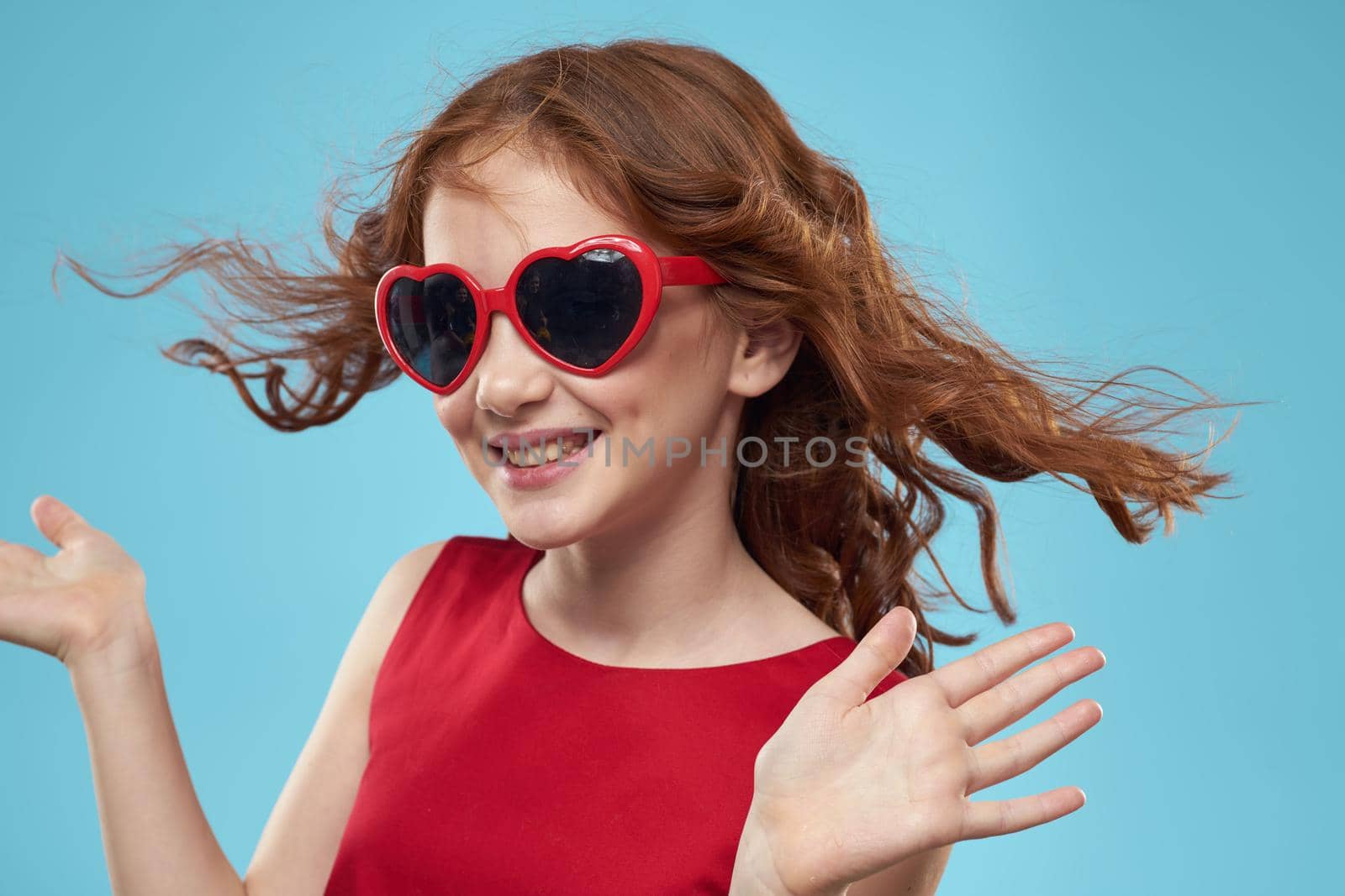 Little girl in sunglasses in the form of hearts red dress curly hair blue background. High quality photo