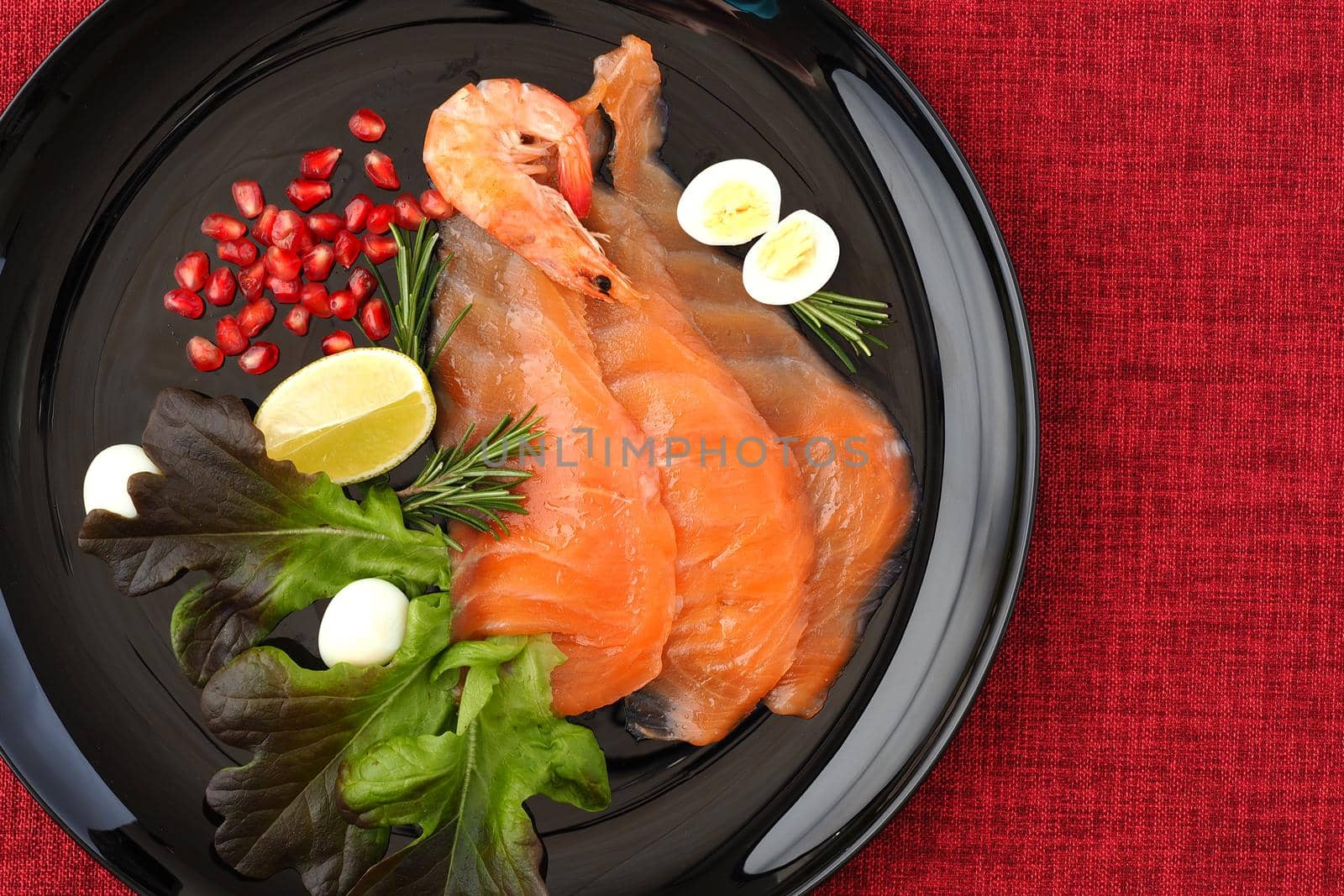 A seafood dish. Sliced salmon slices on a beautiful black plate with lemon, shrimp, quail egg and herbs and a red background. High quality photo