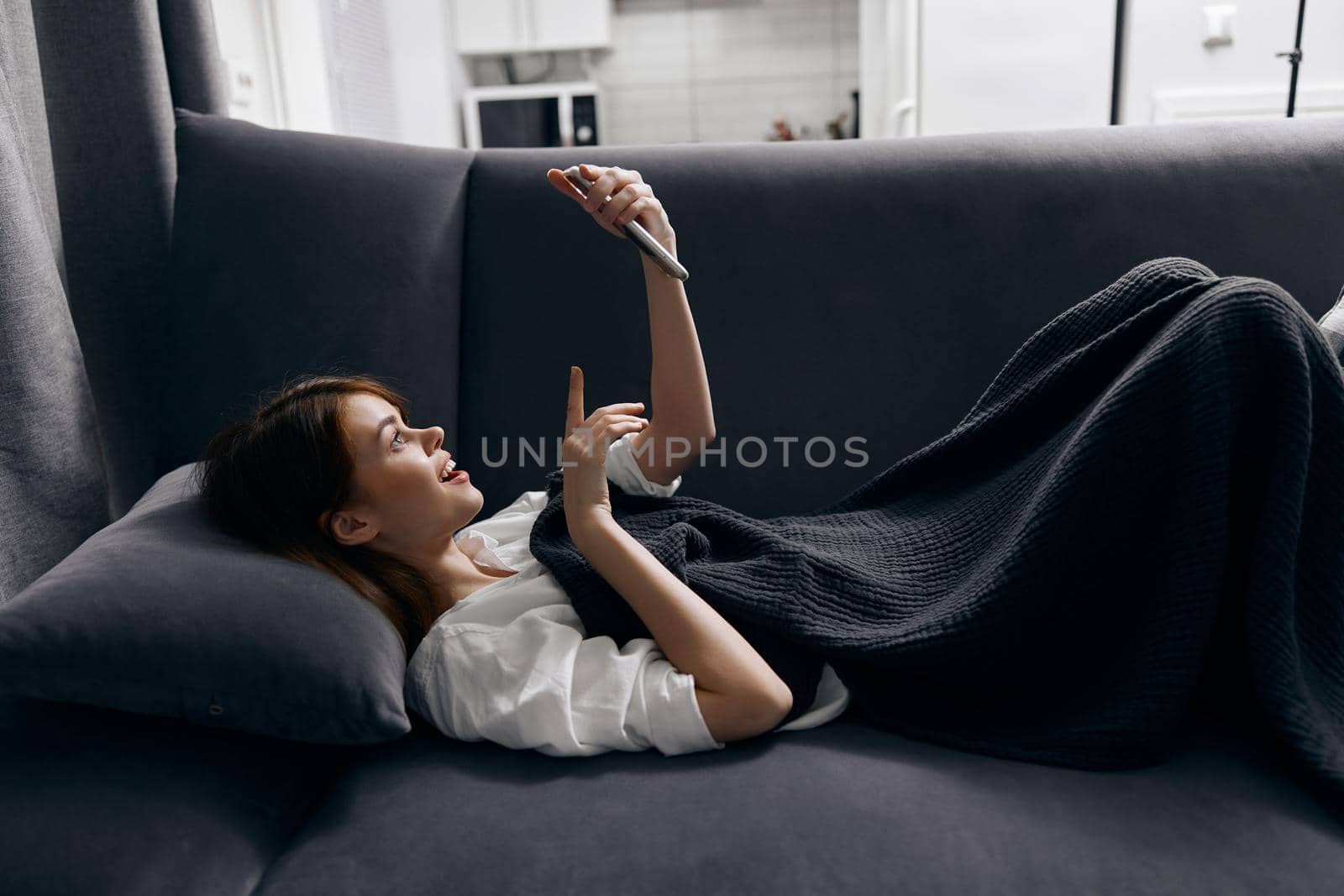 woman looking at the phone screen hiding with a blanket on the sofa. High quality photo