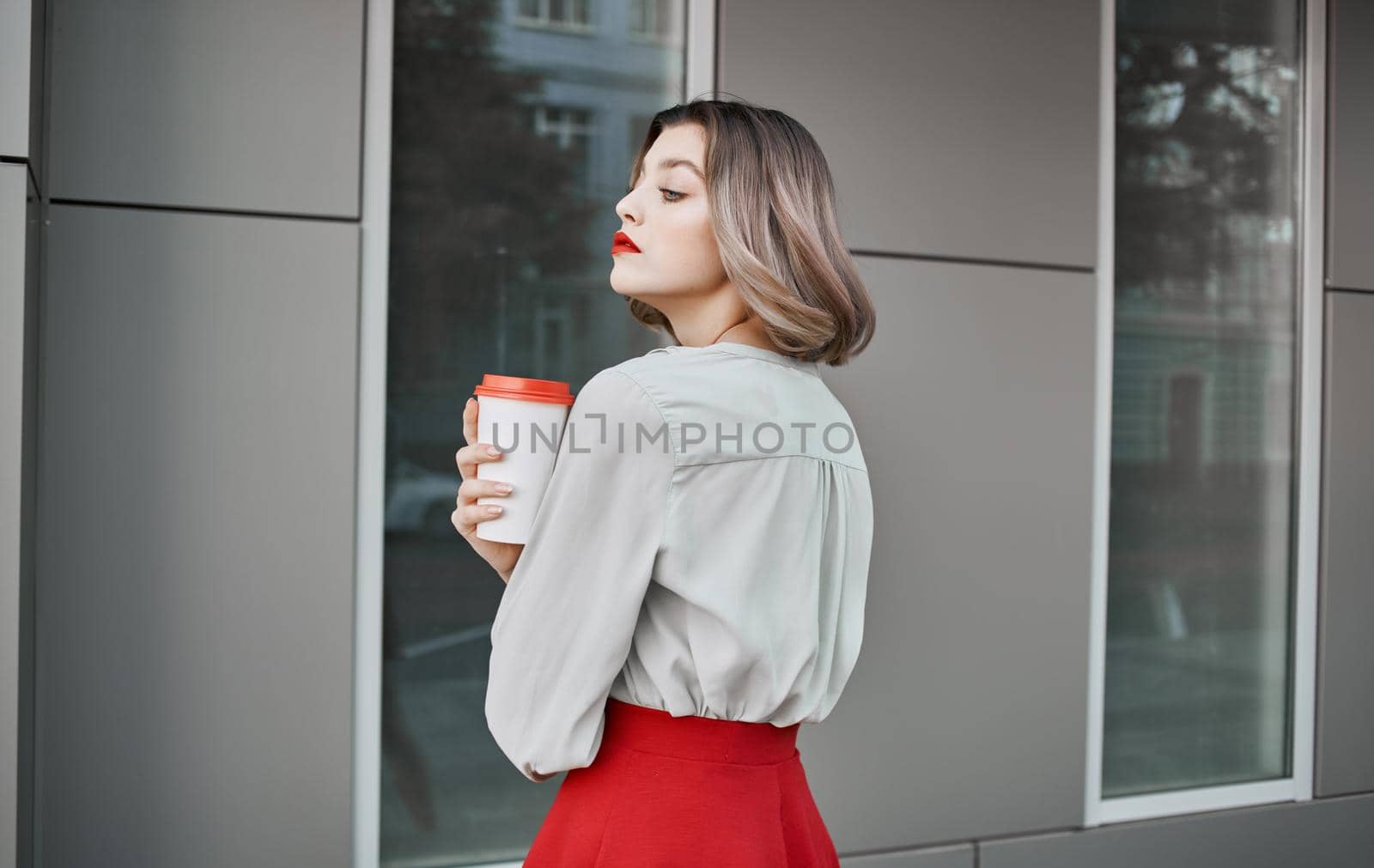 pretty blonde with a cup of coffee near the building on the street and red skirt white shirt. High quality photo