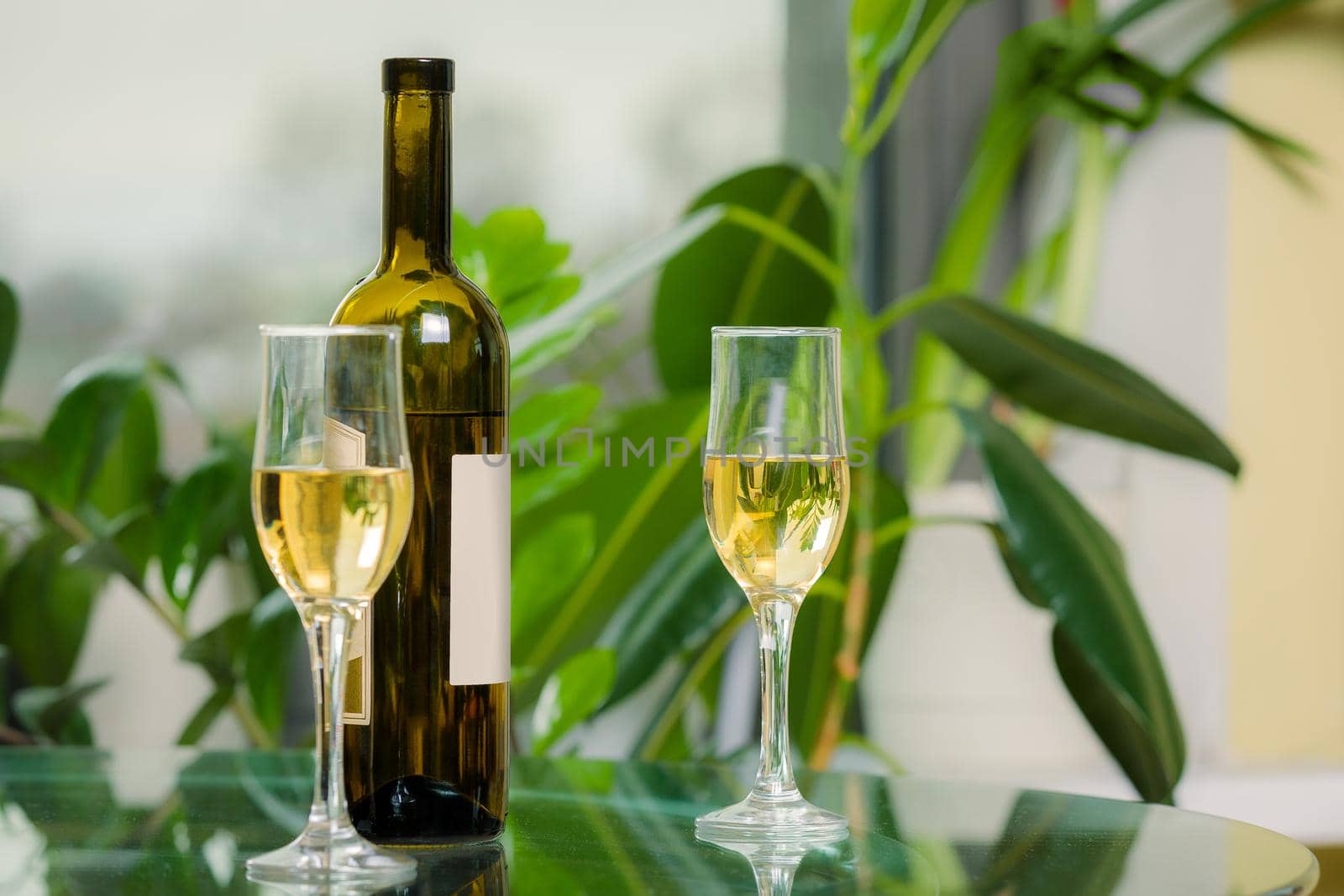 A bottle of white wine with two filled glasses on the background of a window with flowers