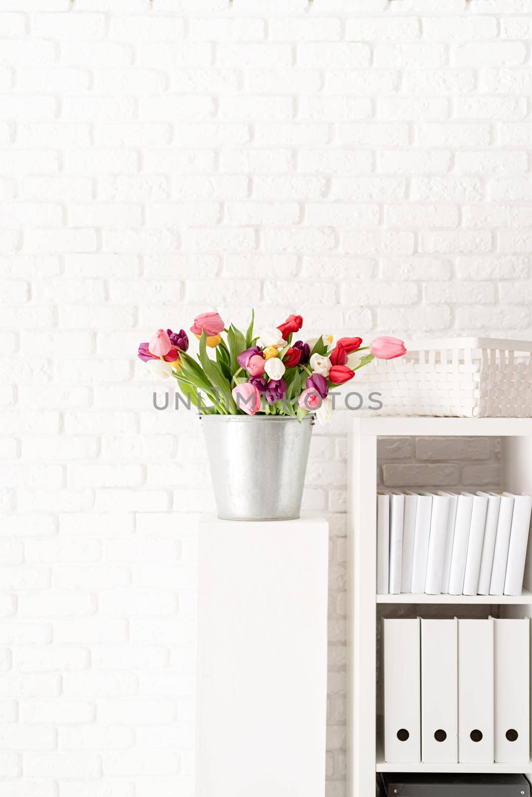 Bucket of tulip flowers next to the bookshelf over white brick wall background by Desperada