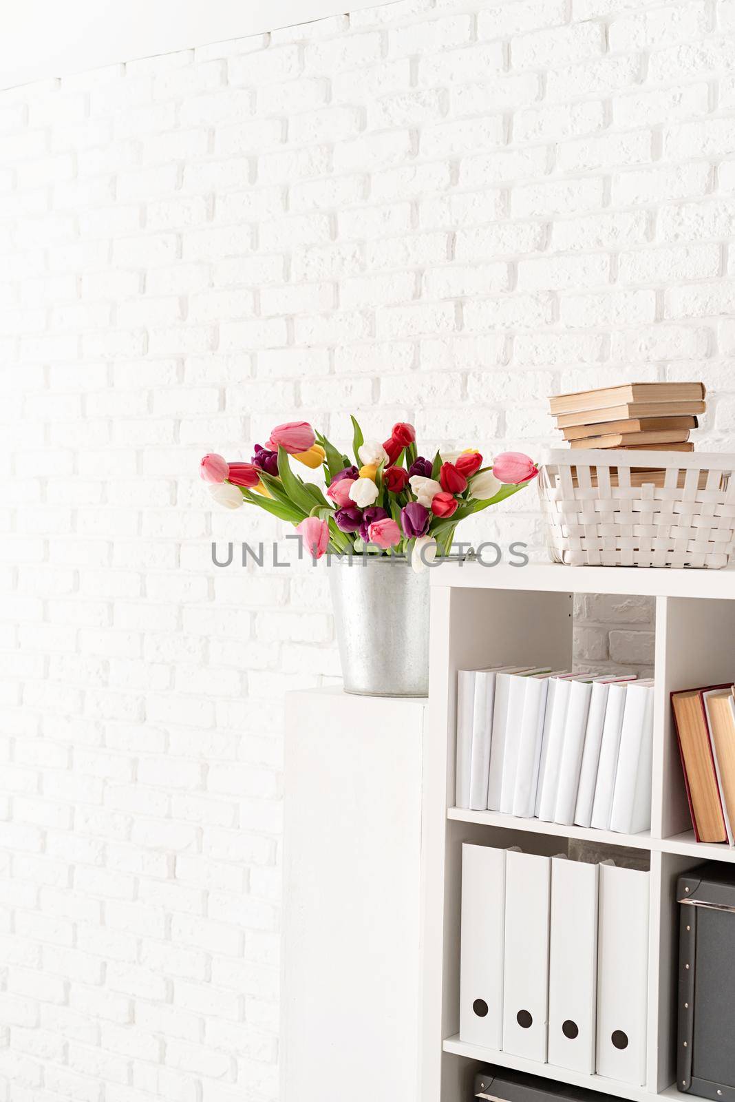Bucket of fresh tulip flowers next to the bookshelf over white brick wall background