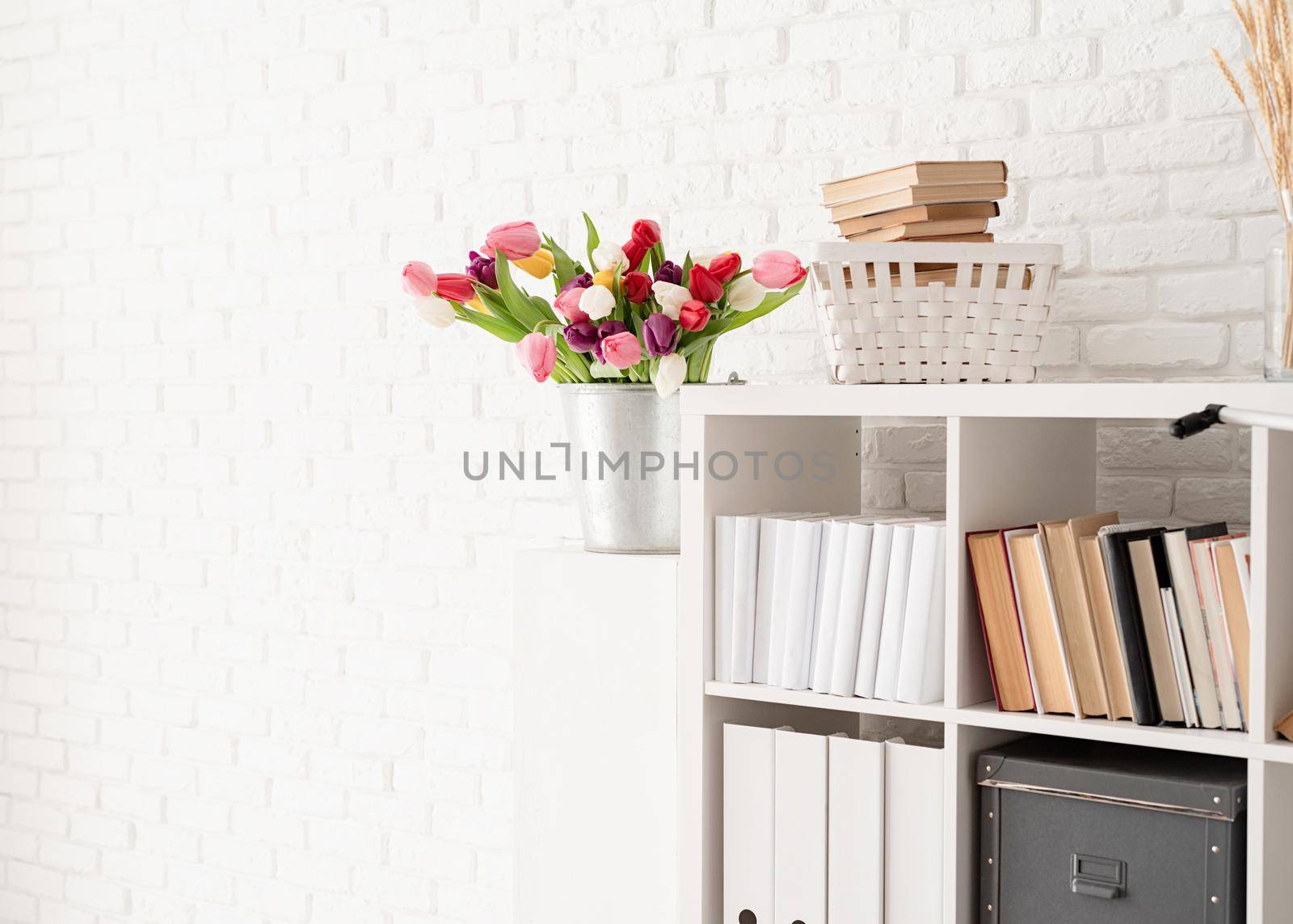 Bucket of tulip flowers next to the bookshelf over white brick wall background by Desperada