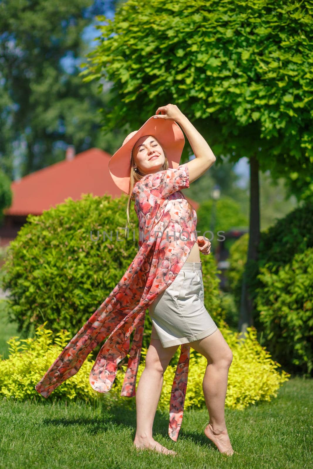 A charming girl in a light summer sundress and a pareo hat is walking in a green park. Enjoys warm sunny summer days.