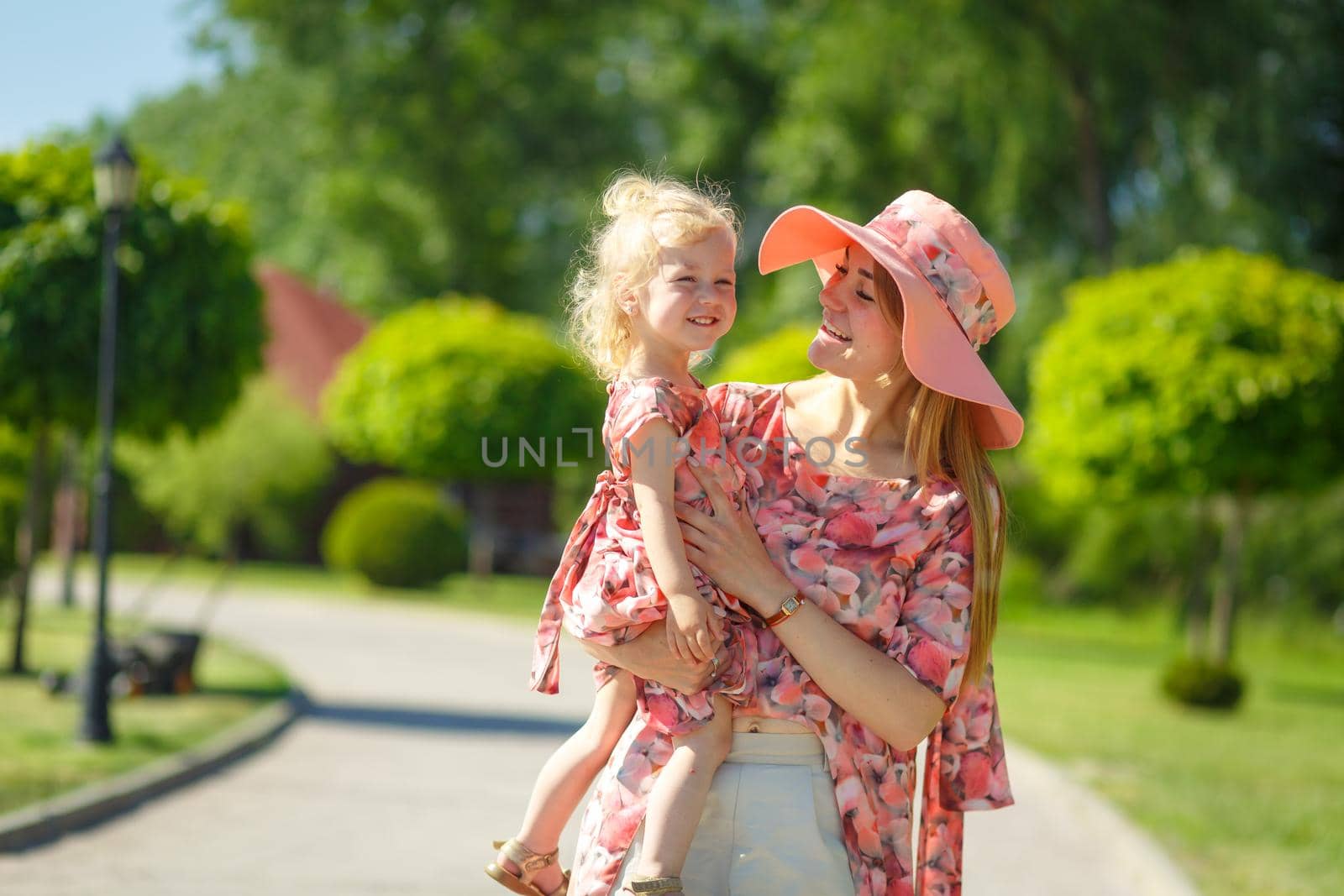 A charming girl in a light summer sundress walks in a green park with her little daughter, holding her in her arms. Enjoys warm sunny summer days.