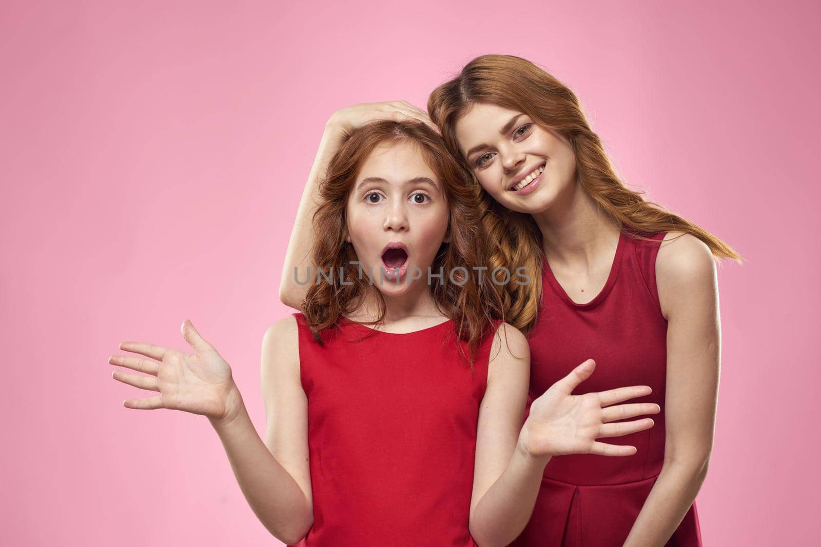 Cheerful mom and daughter wearing red dresses are standing next to joy family on pink background. High quality photo