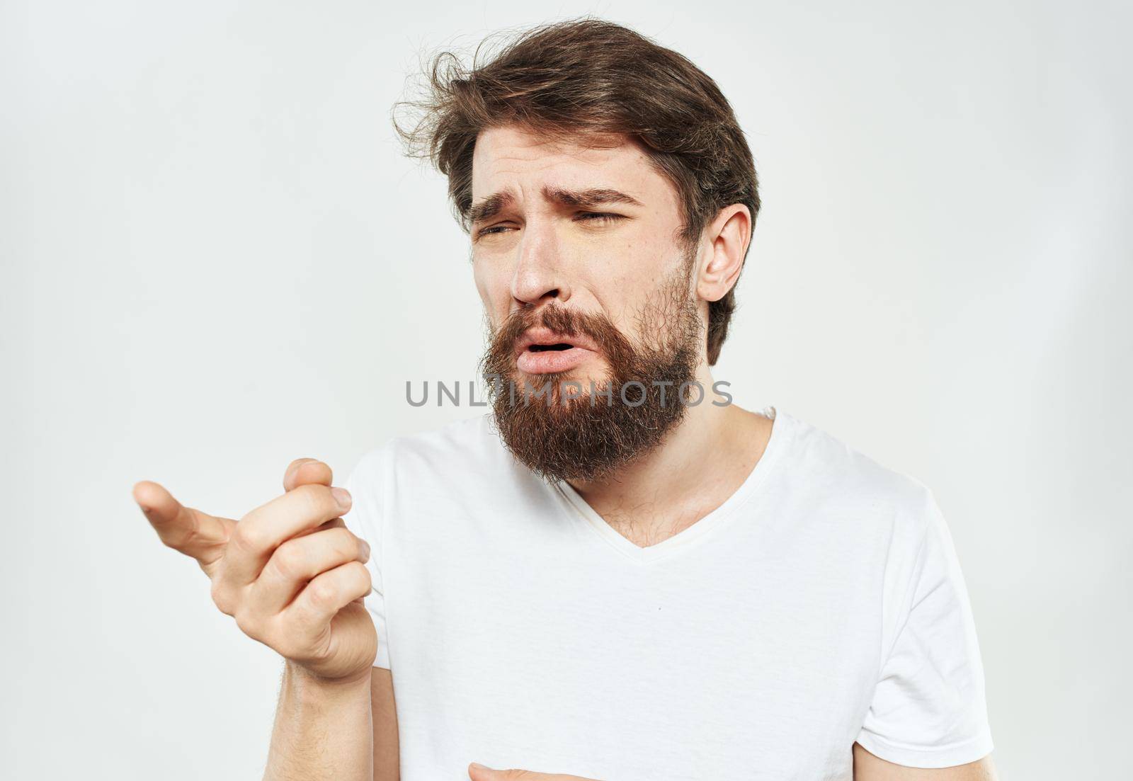 A guy in a white T-shirt with a beard on a light background gestures with his hands emotions model by SHOTPRIME