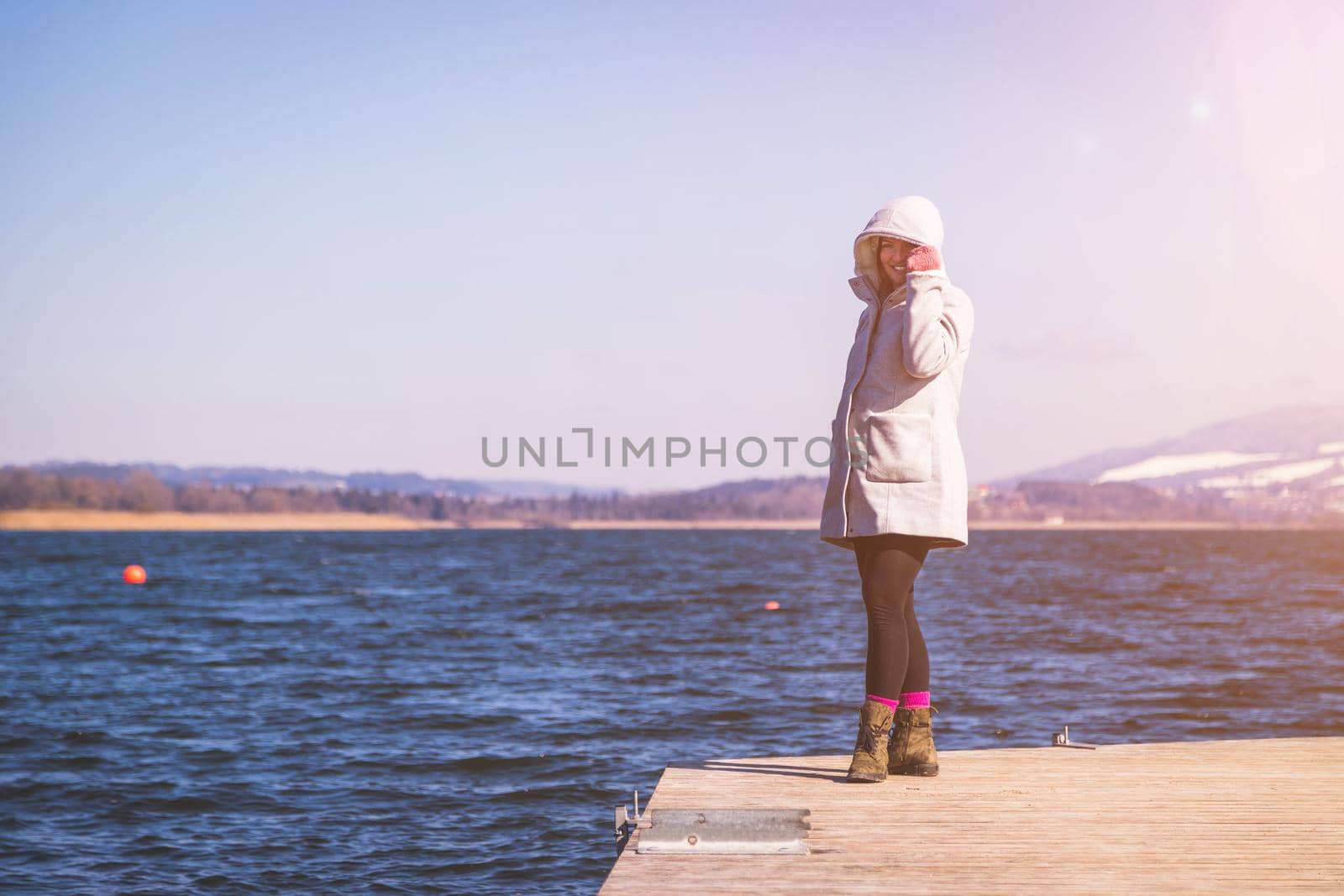Young girl with grey coat is standing on a footbridge and enjoys the view over the lake, winter time
