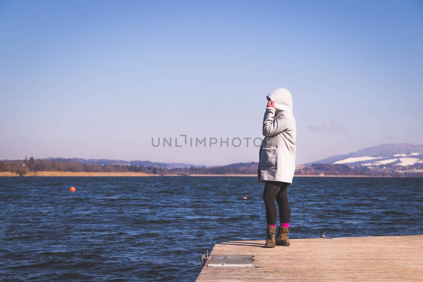 Young girl with grey coat is standing on a footbridge and enjoys the view over the lake, winter time