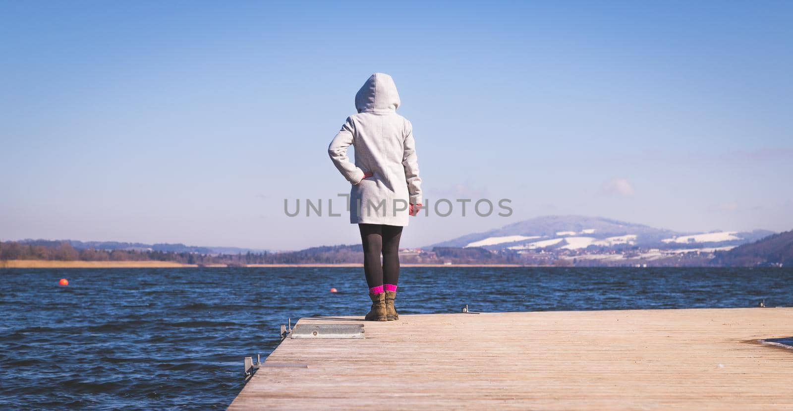 Young girl with grey coat is standing on a footbridge and enjoys the view over the lake, winter time