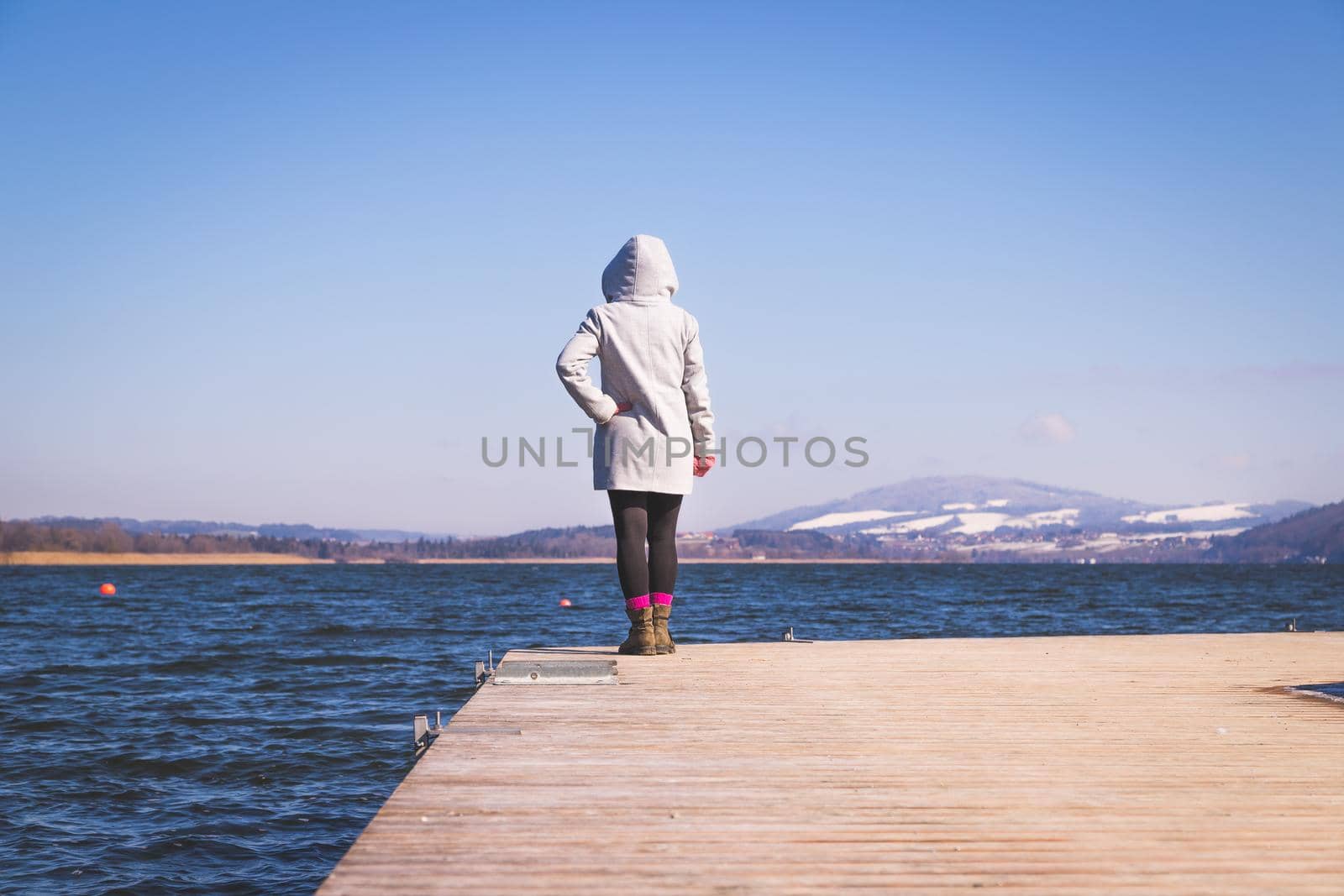 Young girl with grey coat is standing on a footbridge and enjoys the view over the lake, winter time