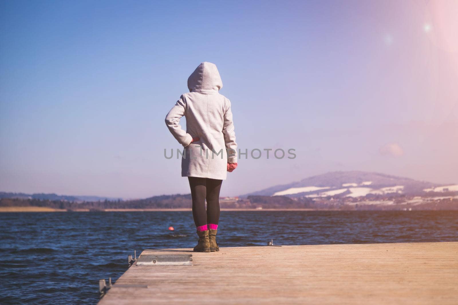 Young girl with grey coat is standing on a footbridge and enjoys the view over the lake, winter time