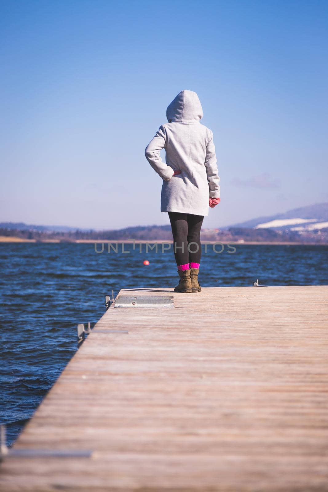 Young girl with grey coat is standing on a footbridge and enjoys the view over the lake, winter time