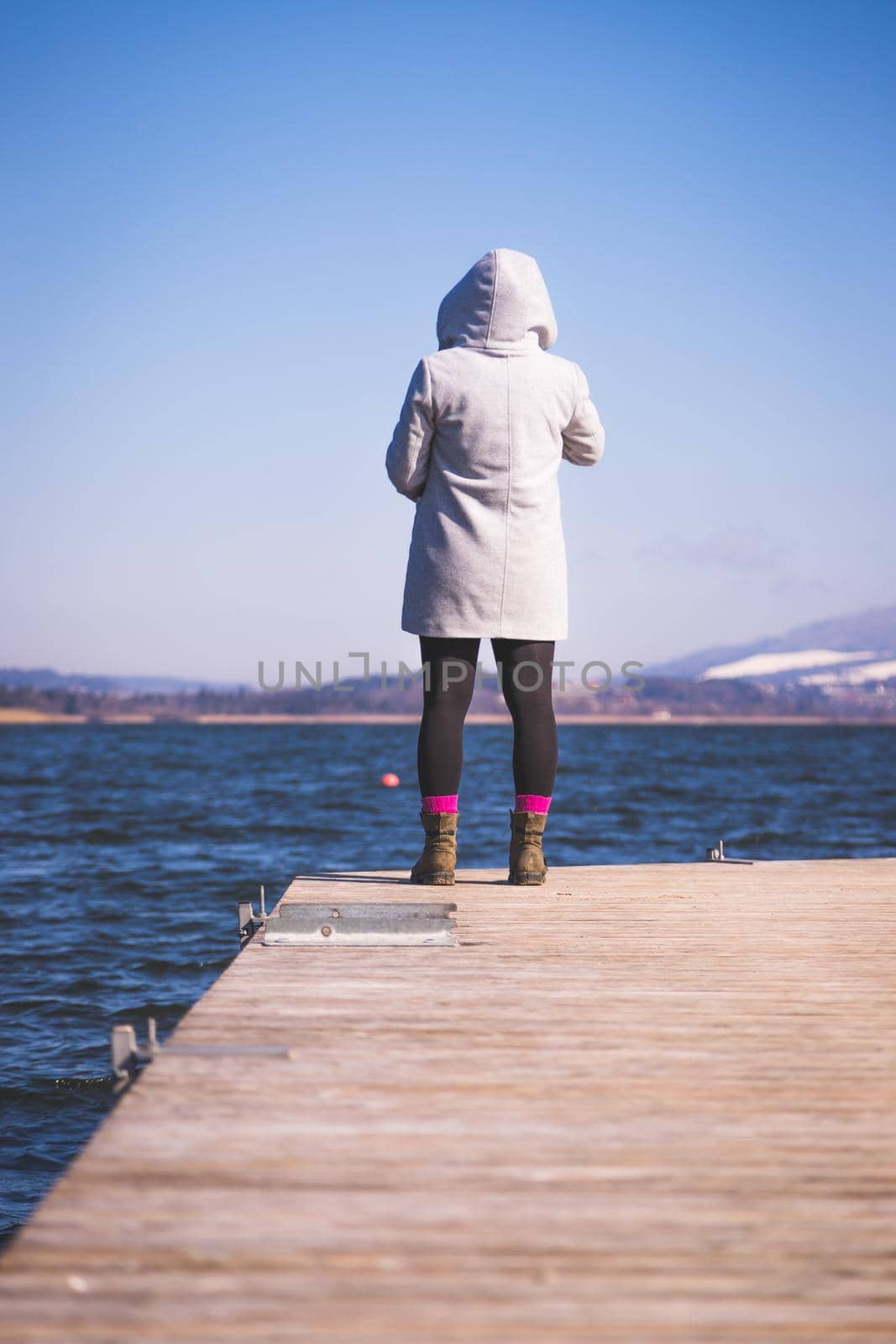 Young girl with grey coat is standing on a footbridge and enjoys the view over the lake, winter time