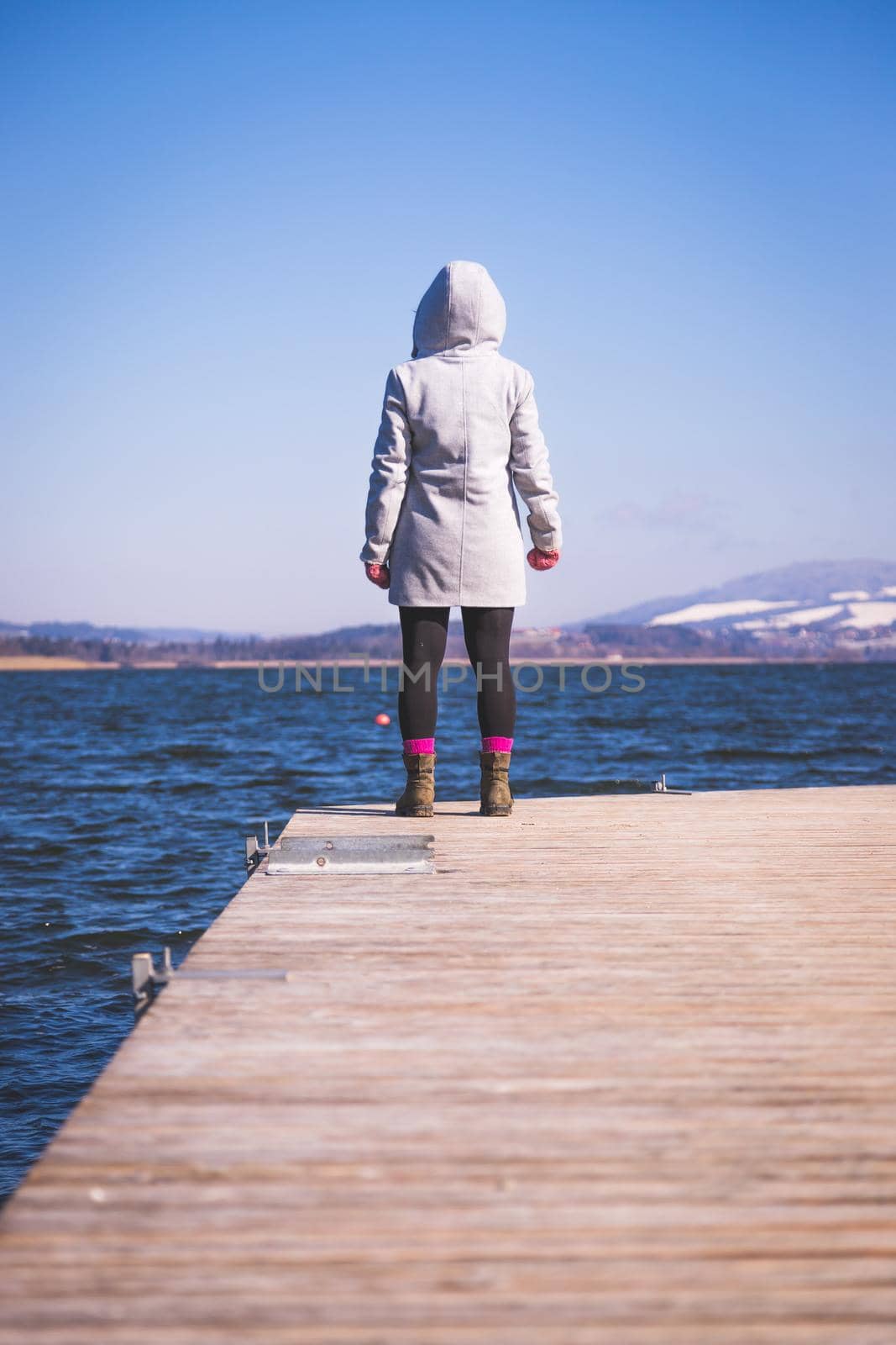 Young girl with grey coat is standing on a footbridge and enjoys the view over the lake, winter time