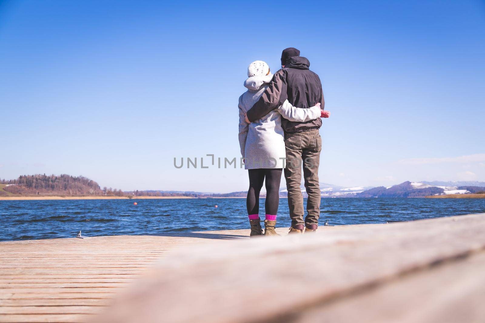 Happy young couple is standing on a footbridge and enjoying the view over the lake, winter time by Daxenbichler