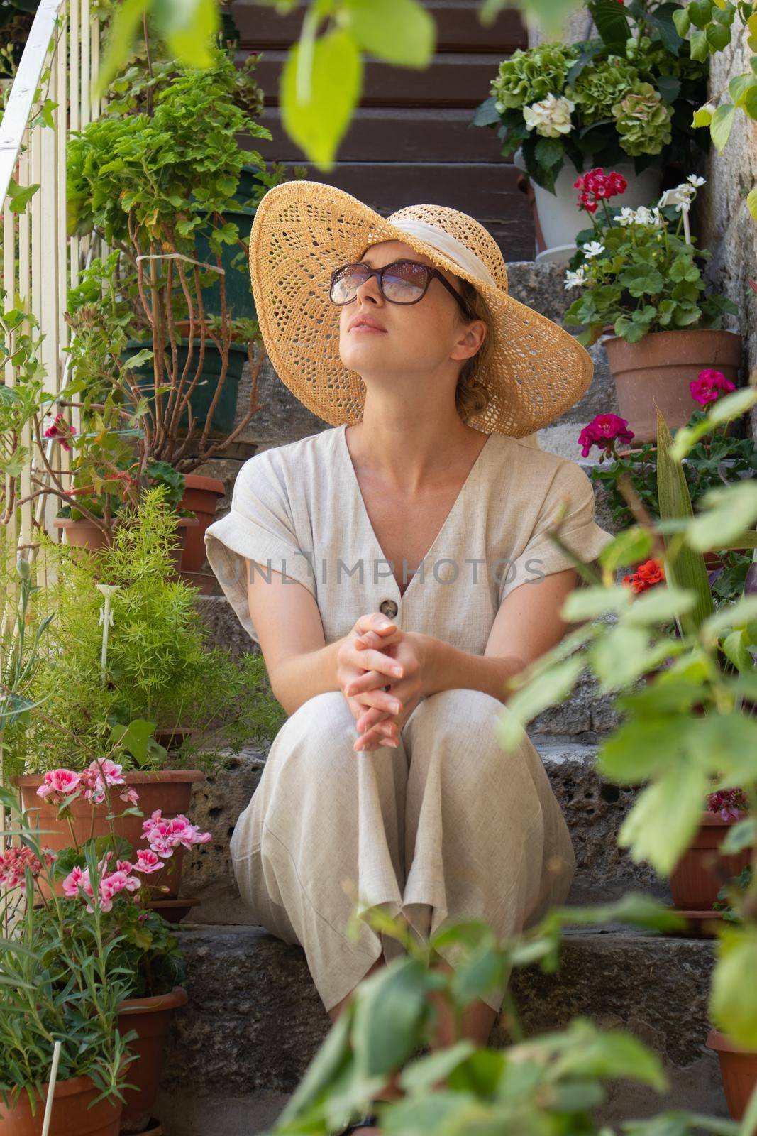 Beautiful female tourist wearing big straw sun hat and sundress sitting and relaxing on old stone house stairs during summer travel on Mediterranean cost on hot summer day