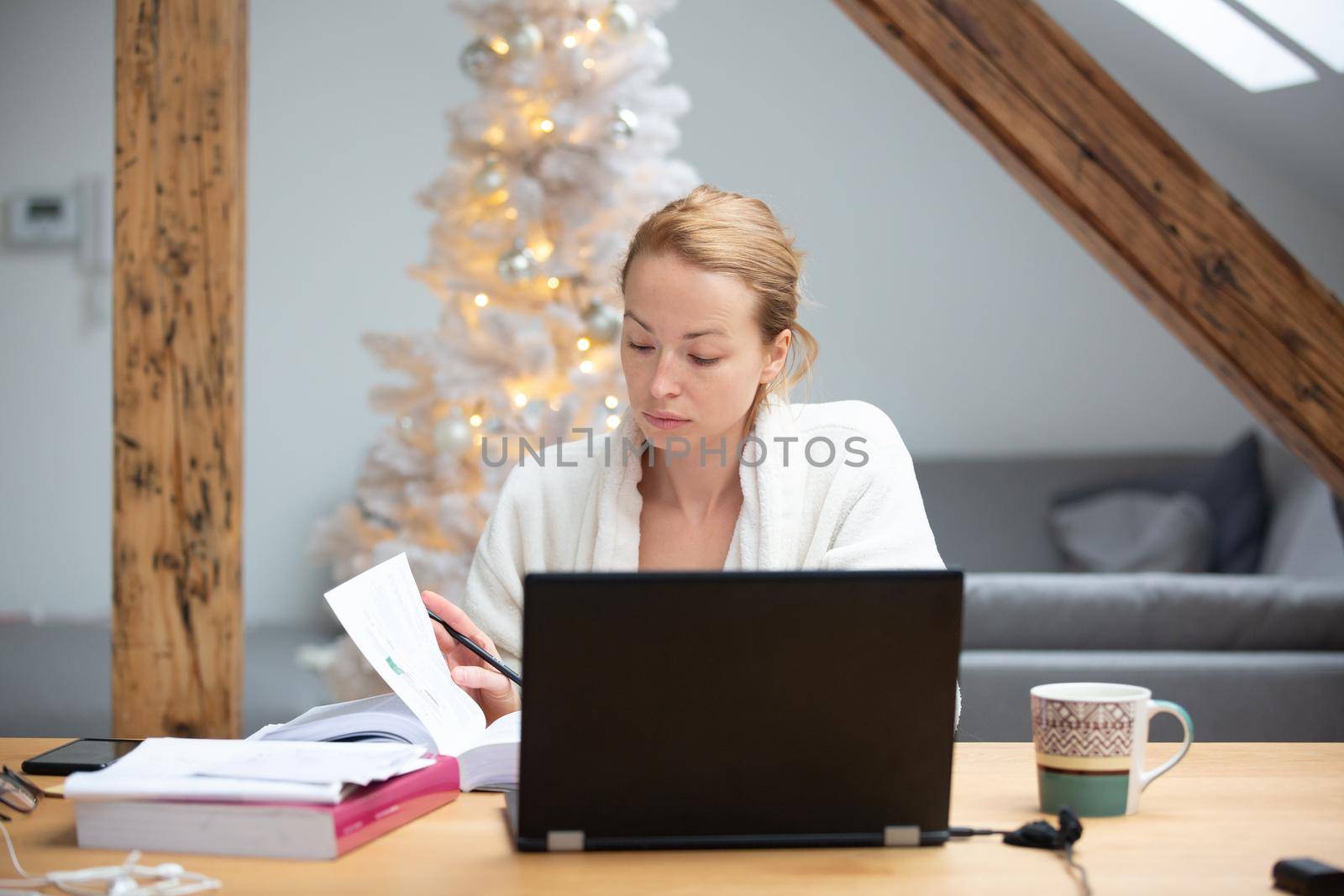 Young businesswoman wearing cosy warm bathrobe working remotly from home in winter Christmas time during 2020 corona virus pandemic. Work from home, selter in place, concept.