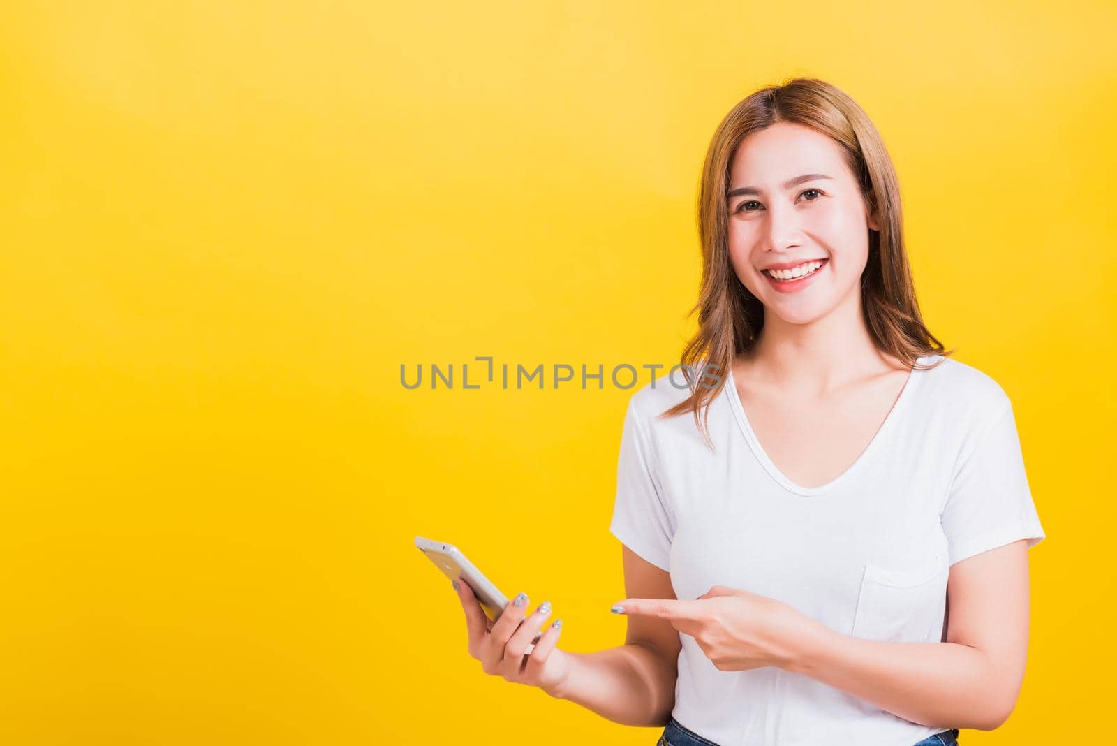 Asian Thai portrait happy beautiful young woman smile standing wear t-shirt making finger pointing on smartphone blank screen looking to camera isolated, studio shot yellow background with copy space