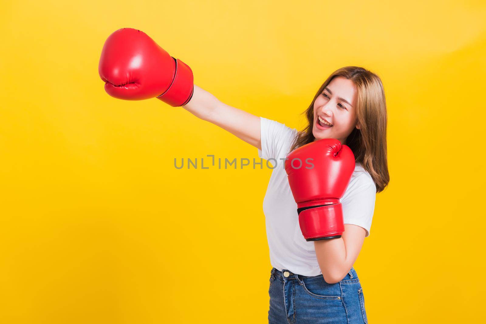 Portrait Asian Thai beautiful young woman standing smile wearing red boxing gloves and punch to side away space, studio shot isolated on yellow background, There was copy space for text