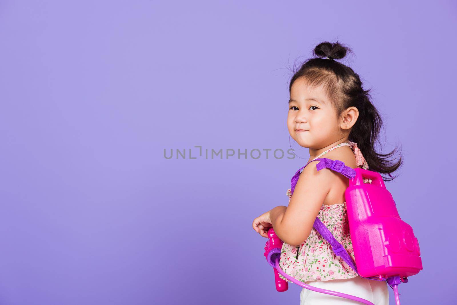 Thai child funny hold toy water pistol and smile, Happy Asian little girl holding plastic water gun, studio shot isolated on purple background, Thailand Songkran festival day national culture concept