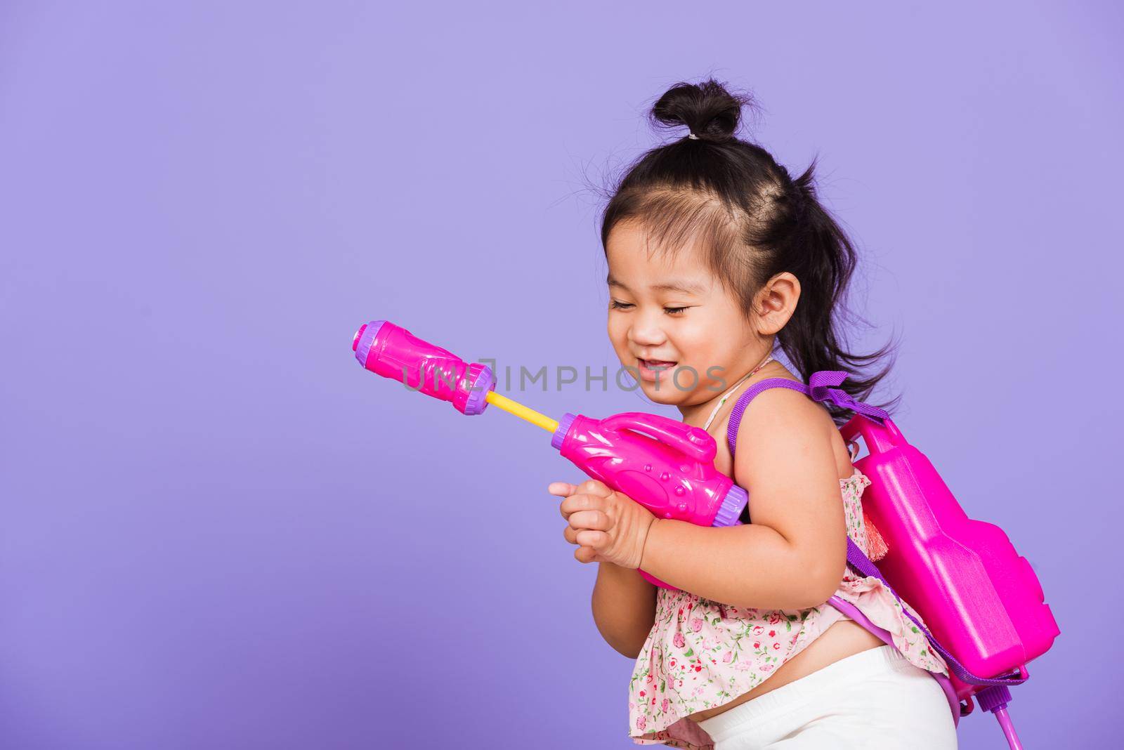 Thai child funny hold toy water pistol and smile, Happy Asian little girl holding plastic water gun, studio shot isolated on purple background, Thailand Songkran festival day national culture concept