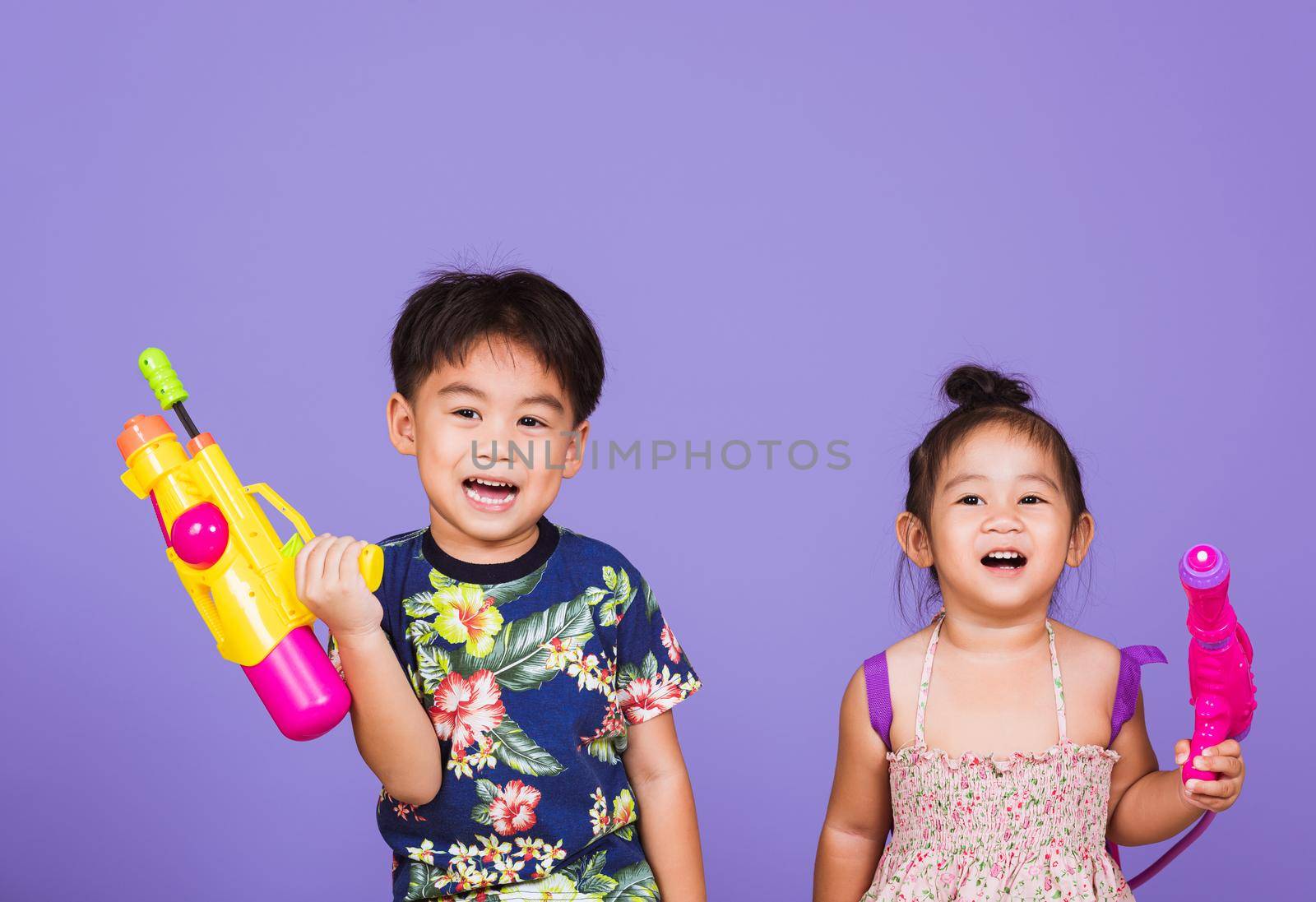 Two Happy Asian little boy and girl holding plastic water gun, Thai children funny hold toy water pistol and smile, studio shot isolated on purple background, Thailand Songkran festival day culture.