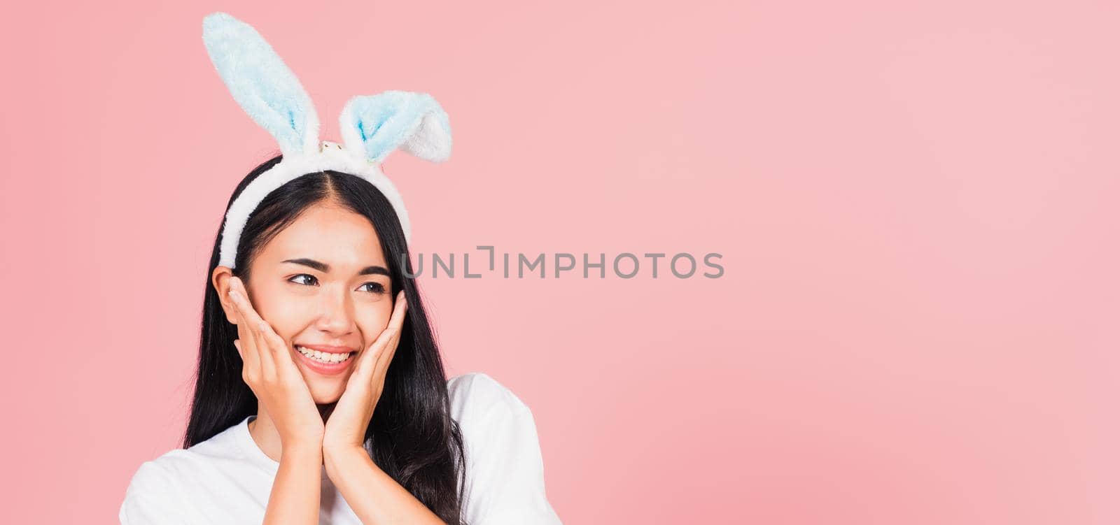 Happy Easter Day. Beautiful young woman teen smiling wearing Easter rabbit bunny ears holding her cheeks excited surprised, Portrait female face touch massage, studio shot isolated on pink background