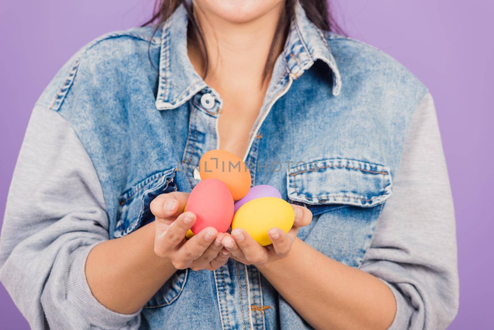 Happy Easter concept. Beautiful young woman smiling wearing rabbit ears and denims hold colorful Easter eggs gift on hands, Portrait female looking at egg, studio shot isolated on purple background