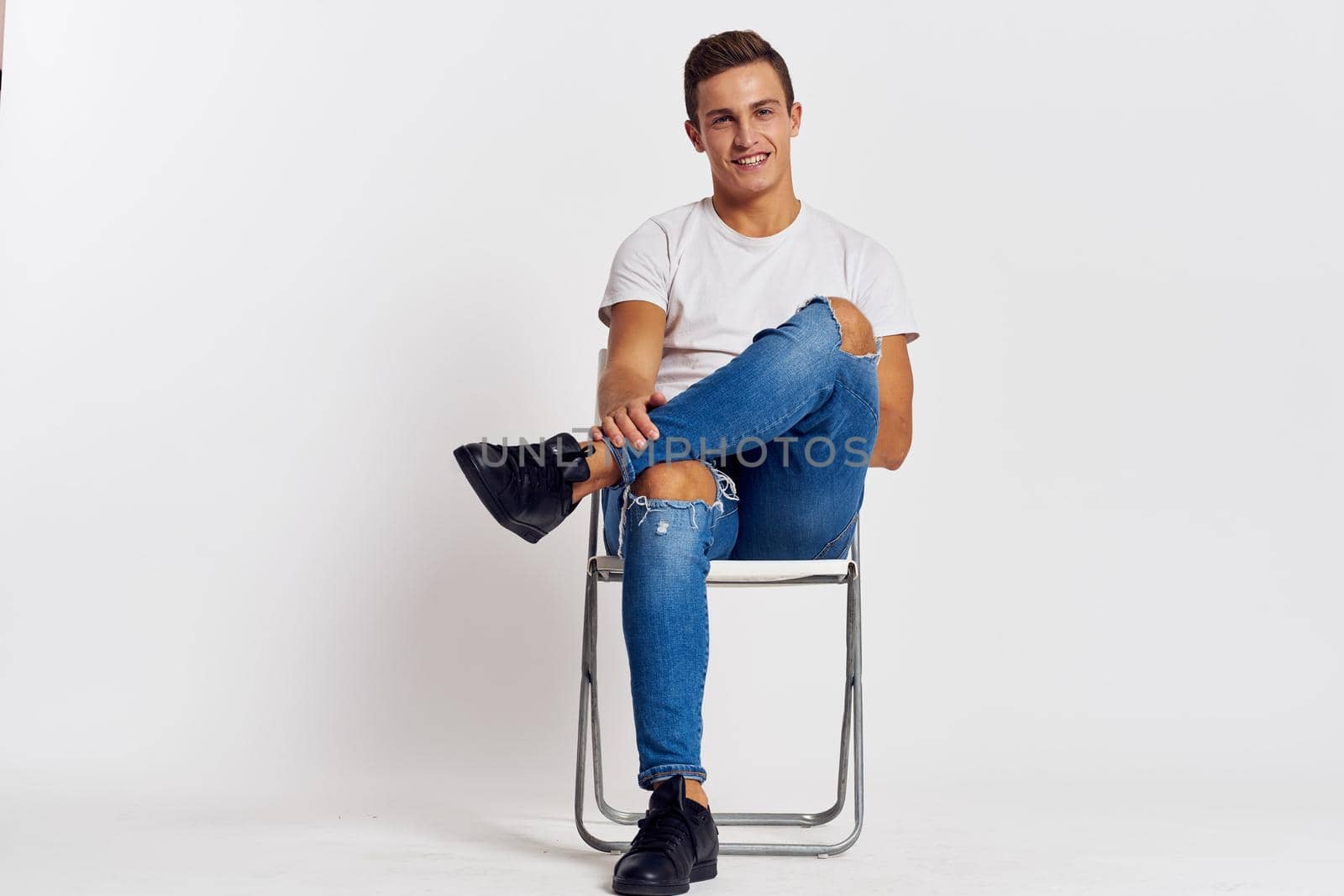 Man on a chair indoors torn jeans white t-shirt handsome face model light background by SHOTPRIME
