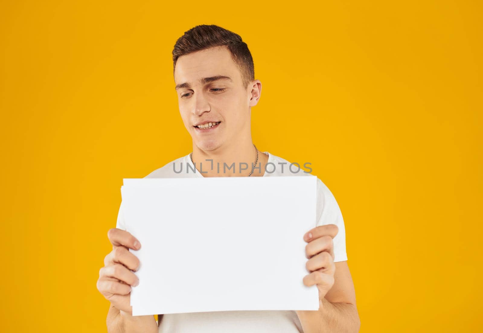 A man in a white T-shirt with a sheet of paper on a yellow background Copy Space mockup by SHOTPRIME