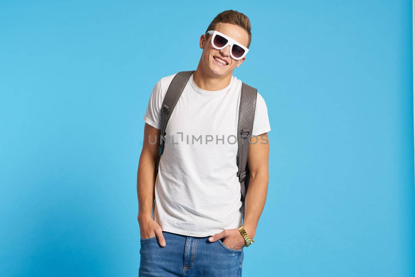 Relaxed guy in jeans with torn knees sits on a chair indoors on a blue background with full-length glasses on his face. High quality photo