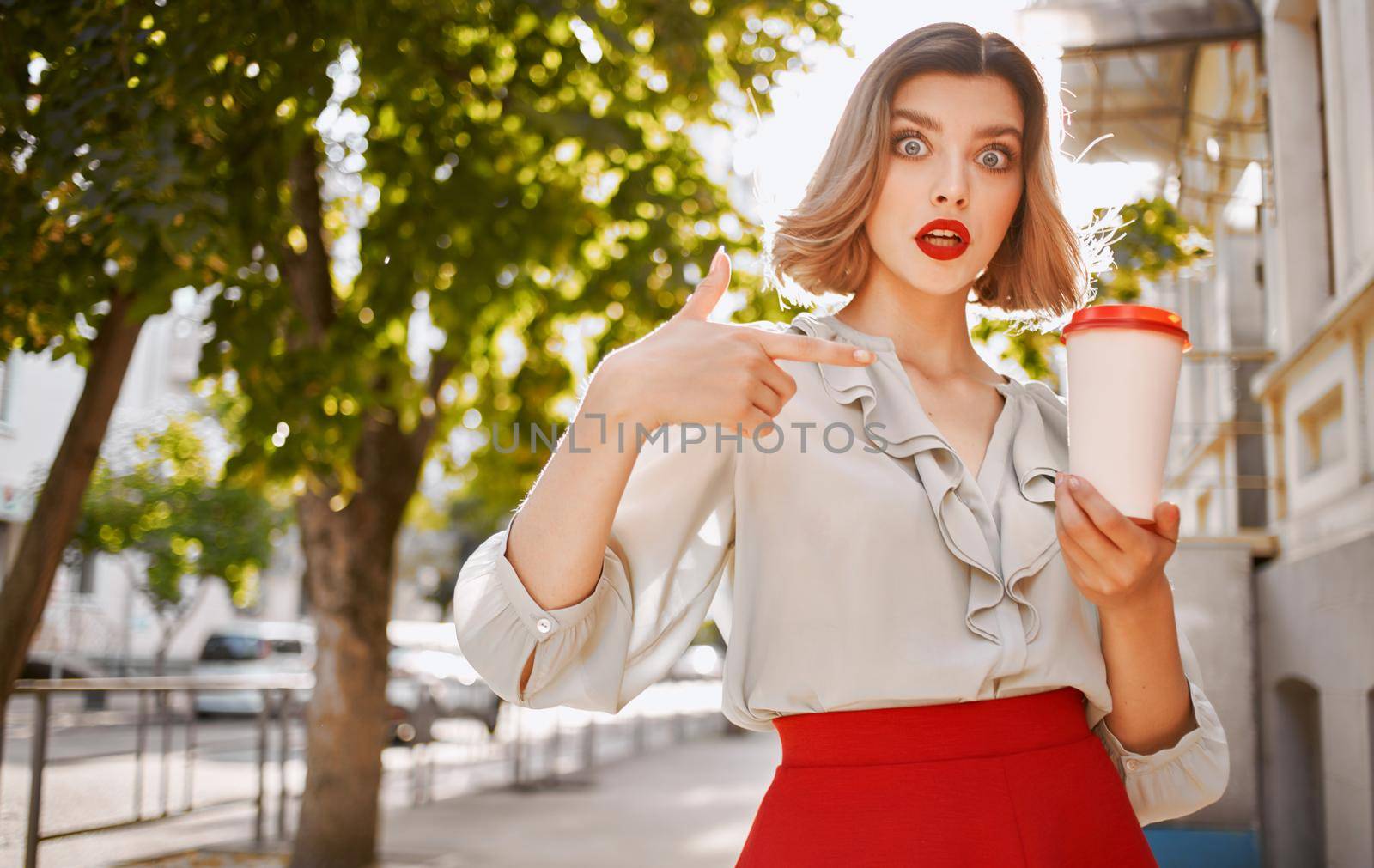 fashionable blonde woman in red skirt stands near the building sidewalk park nature. High quality photo
