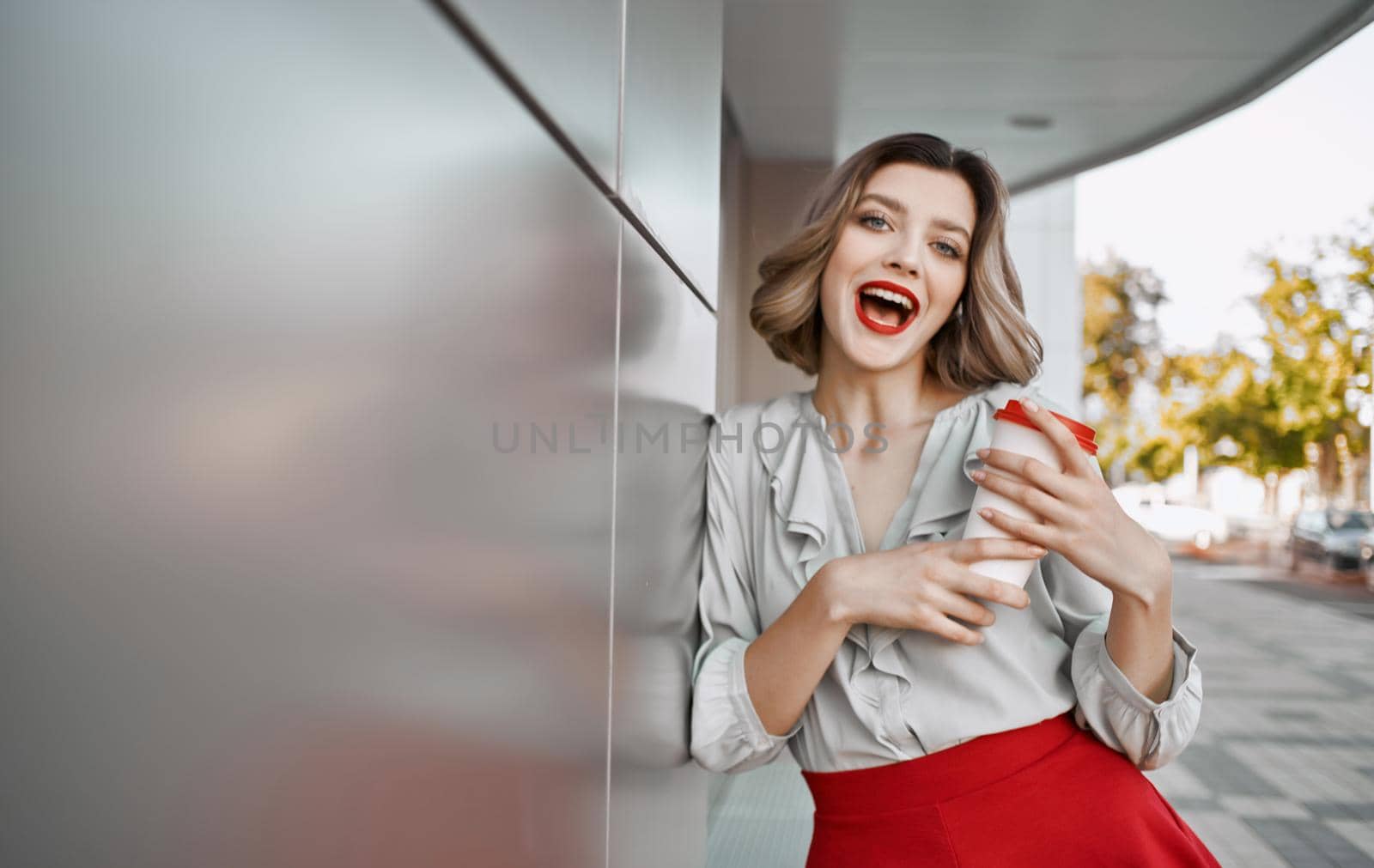 pretty blonde with a cup of coffee near the building on the street and red skirt white shirt. High quality photo