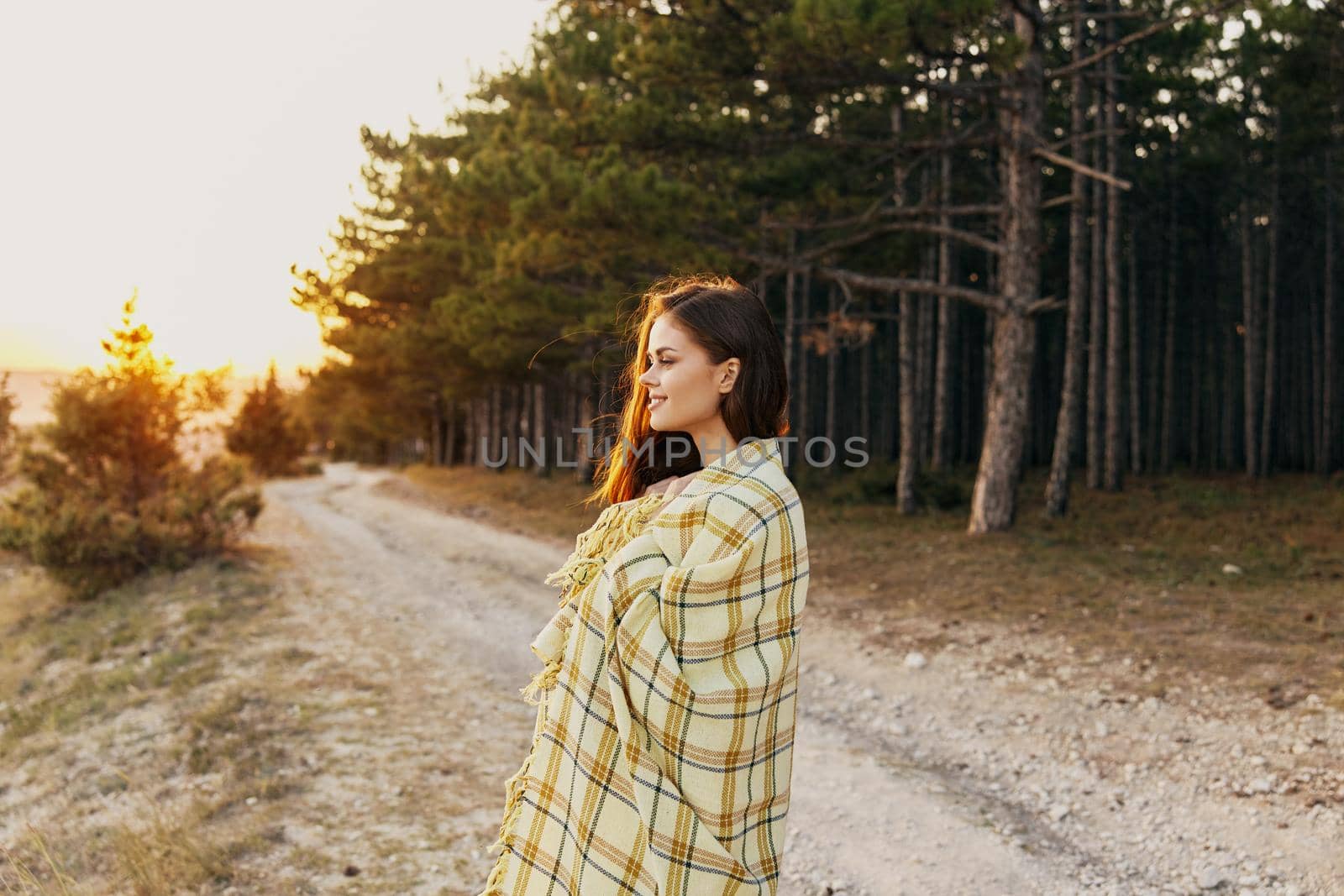 Happy woman tourist with a plaid on her shoulders stands on the road near high fir trees. High quality photo