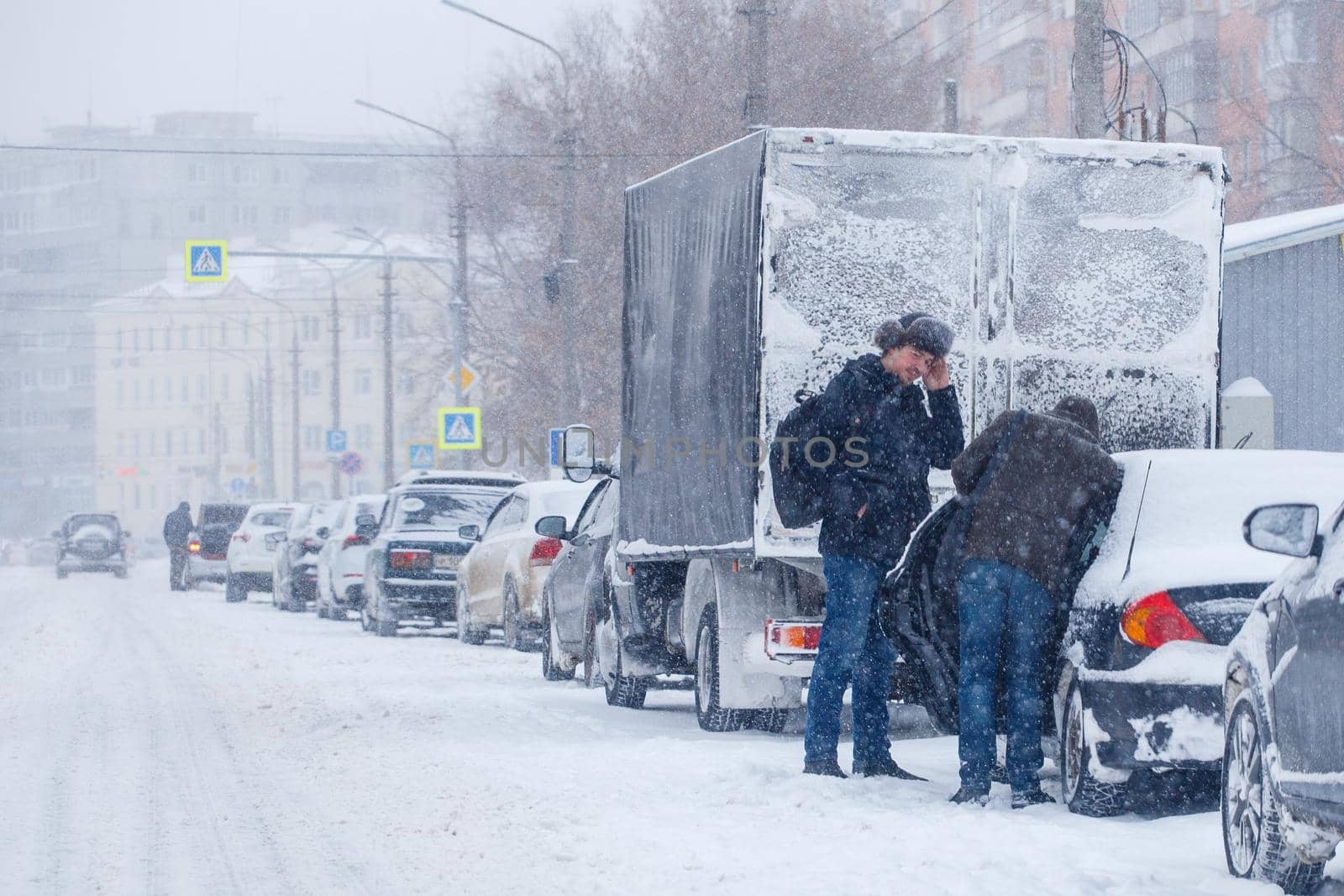 Tula, Russia - February 13, 2020: Two men standin near car parked on side of city road during heavy snowstorm at day light by z1b