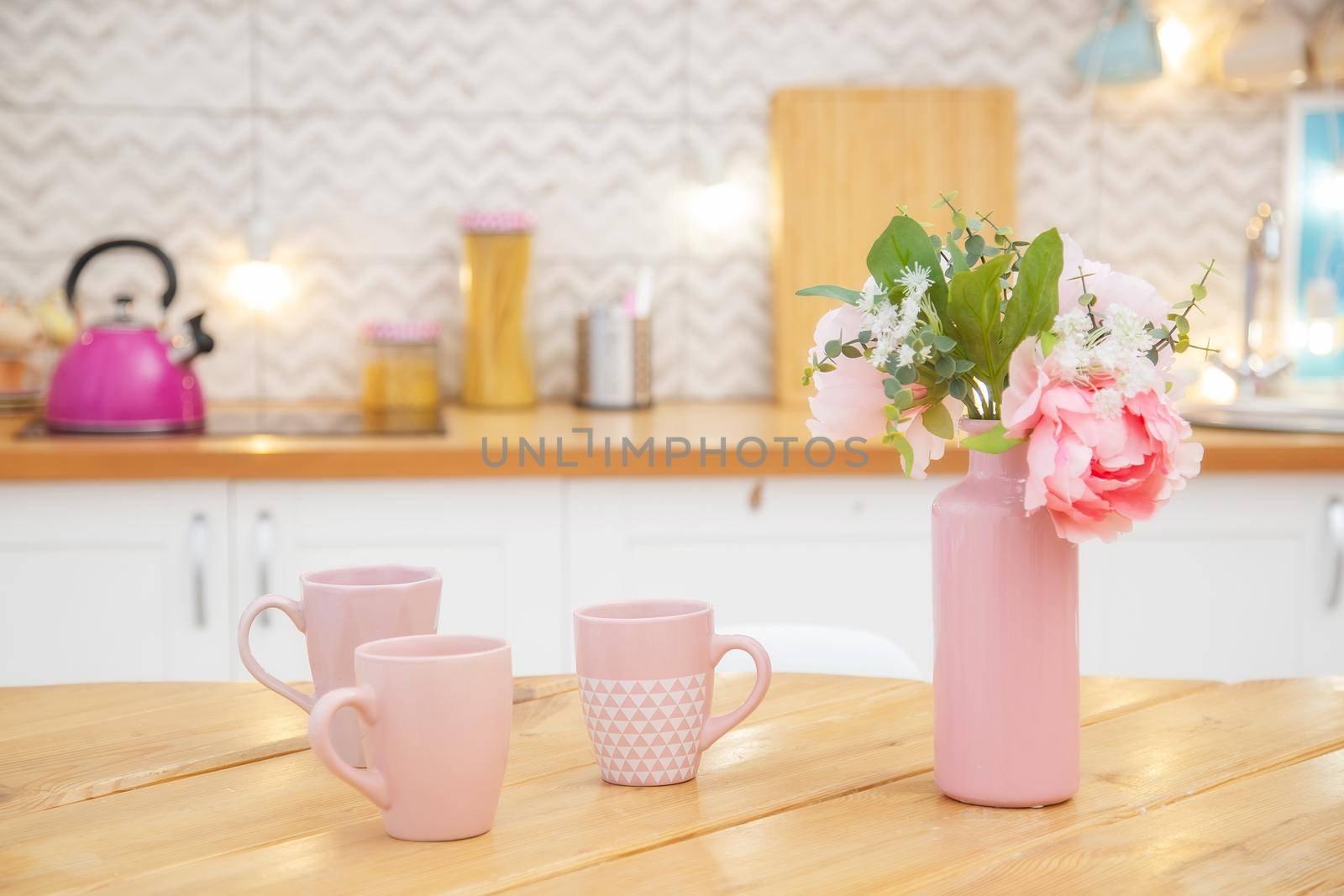 Pink vase with flowers and mugs for tea on the table in a light kitchen in the Scandinavian style.