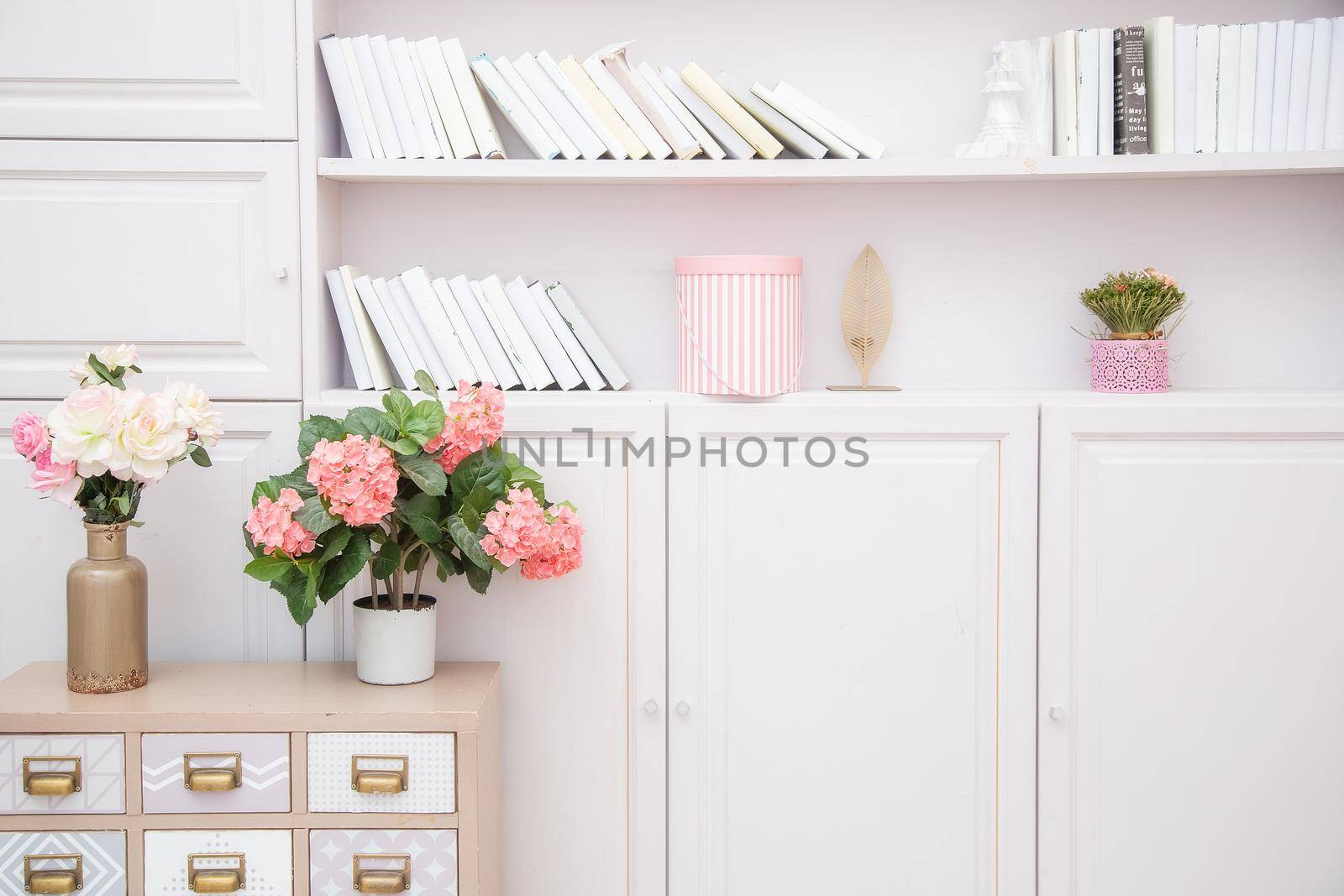 A vase of flowers on a chest of drawers near a bookcase in a light pink living room in a Scandinavian style.