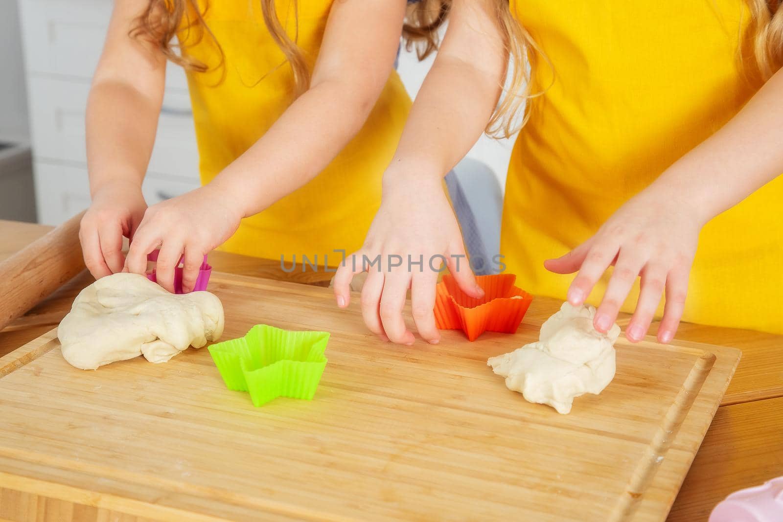 Two sisters of Caucasian appearance prepare cookies in the kitchen using a wooden cutting board, rolling pin and molds. Cropped frame of children in yellow aprons.